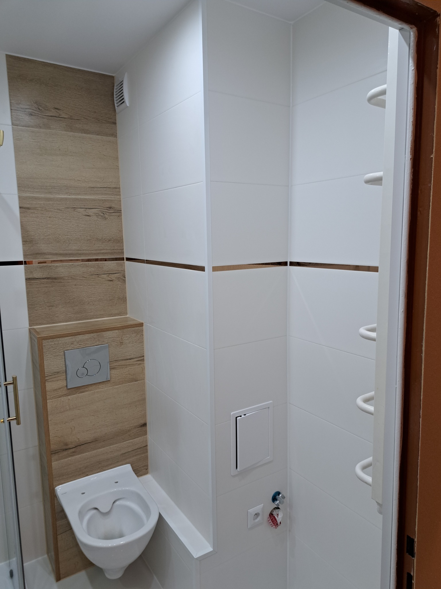 Nowoczesna łazienka po remoncie: białe płytki z poziomymi paskami w kolorze złota, zabudowa WC z drewnopodobną okładziną, wisząca miska WC bez deski, biały grzejnik drabinkowy, wentylator...