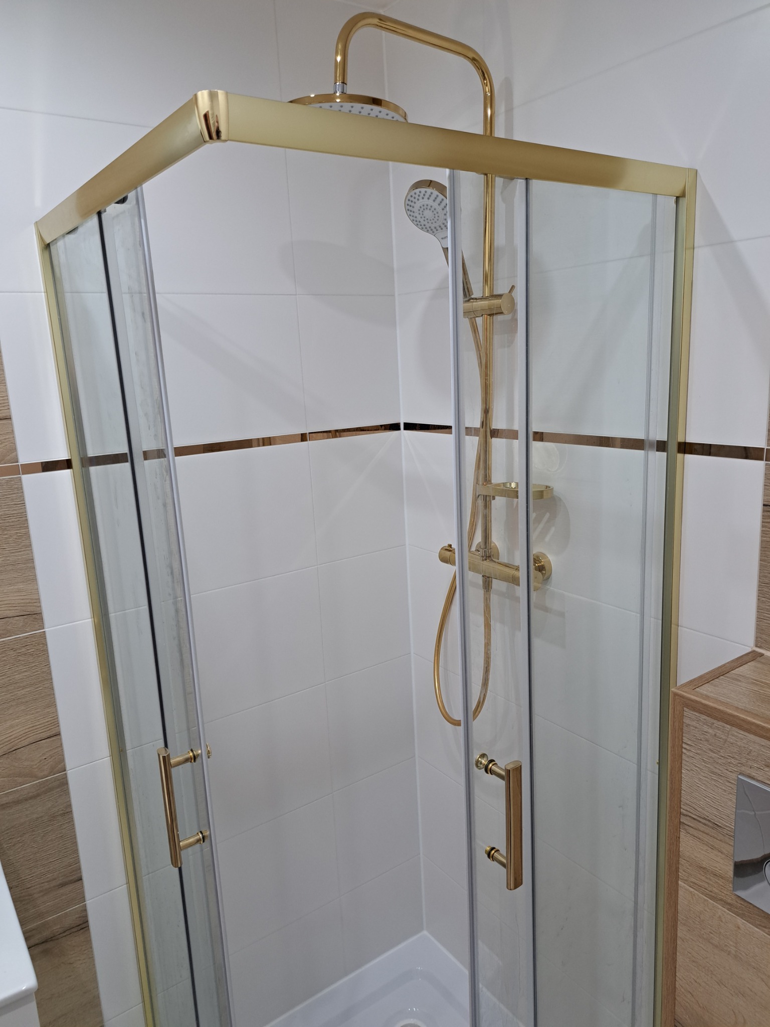 Szklana kabina prysznicowa z złotymi profilami i armaturą w wykończonej łazience z białymi płytkami i drewnopodobnymi akcentami.