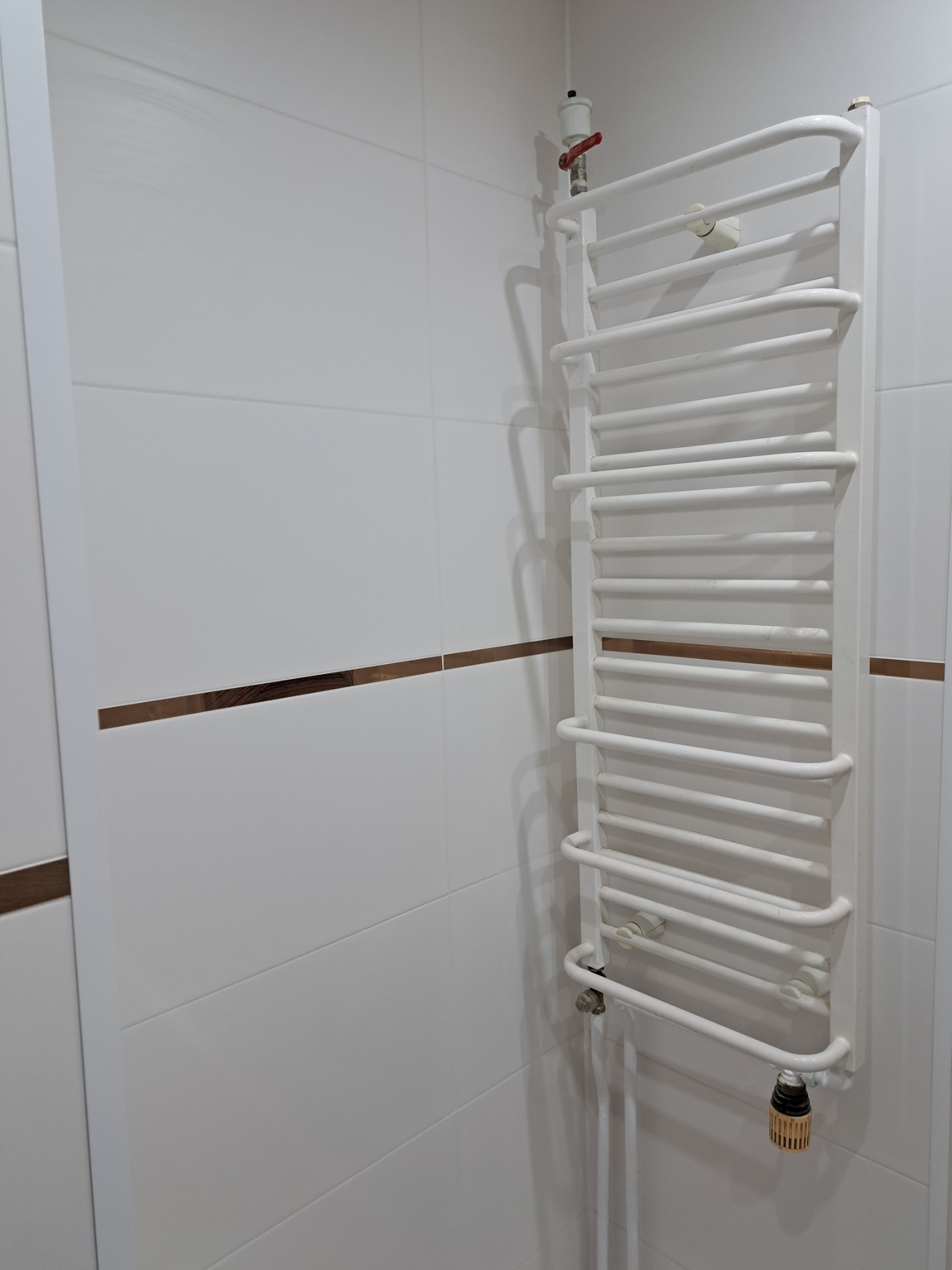 Biały, drabinkowy grzejnik łazienkowy zamontowany na ścianie z białych płytek z brązowym akcentem.