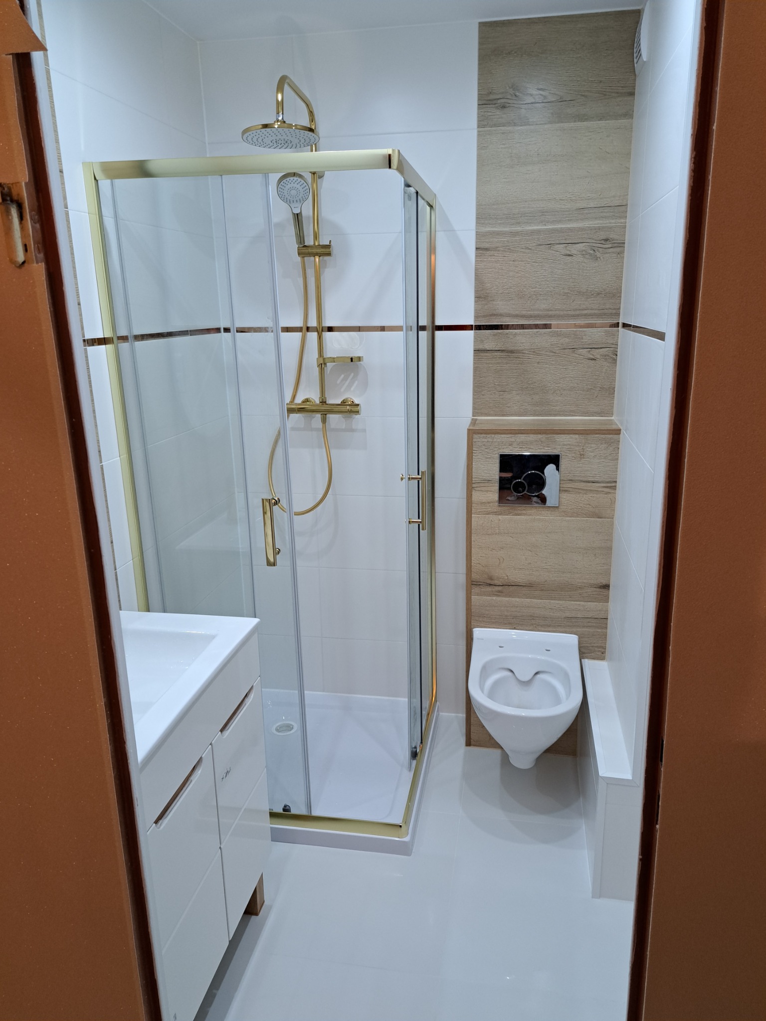 Nowoczesna łazienka z kabiną prysznicową ze złotymi elementami, białą armaturą i płytkami imitującymi drewno na ścianie.