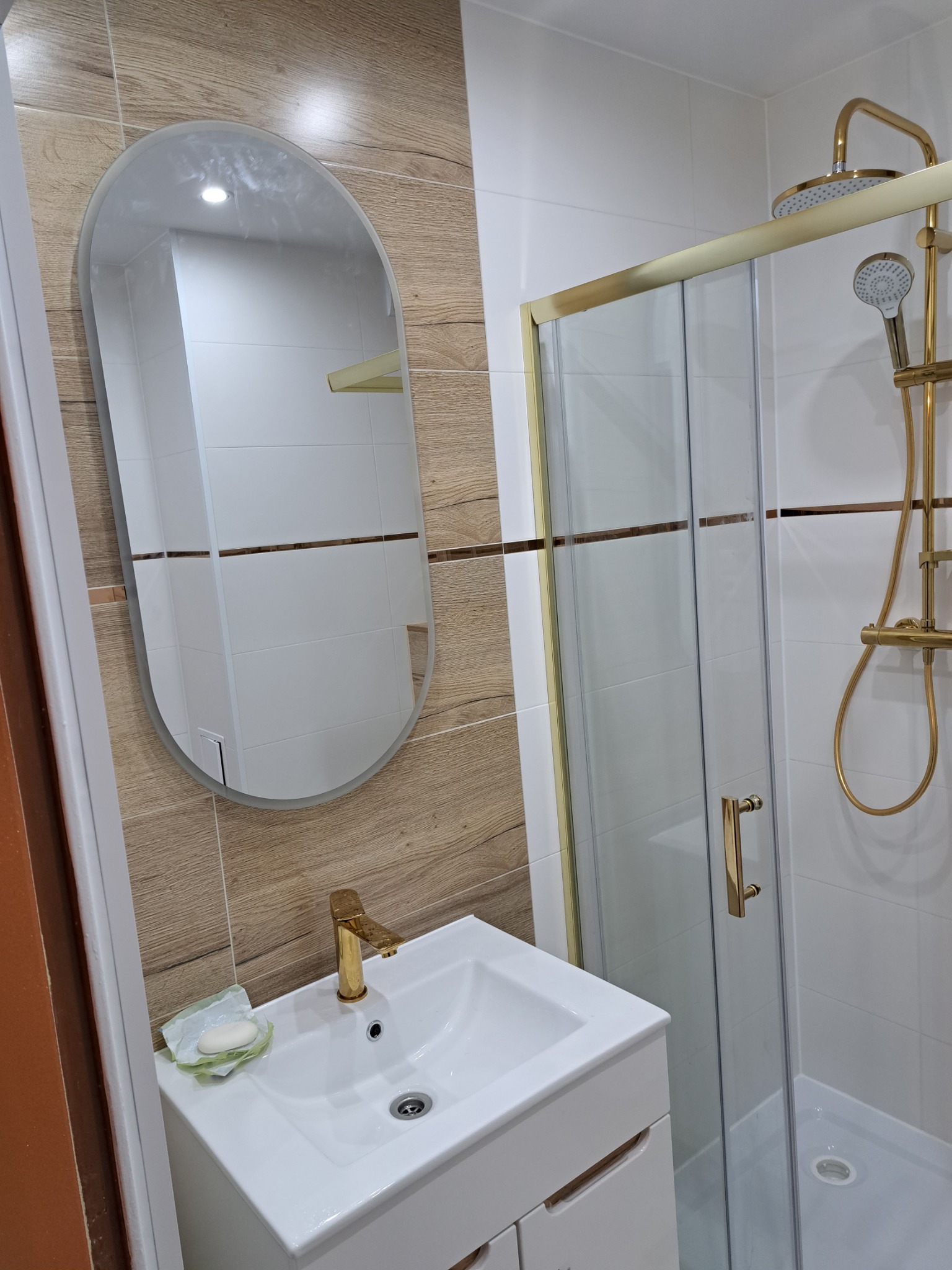 Nowoczesna łazienka z kabiną prysznicową ze złotymi detalami, umywalką z szafką i owalnym lustrem na ścianie wyłożonej płytkami imitującymi drewno.