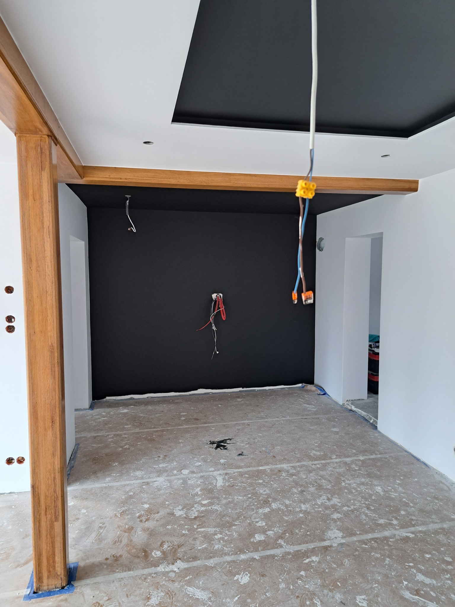 Pokój w trakcie malowania: czarna ściana i sufit kontrastują z białymi ścianami, drewniane elementy dekoracyjne, zabezpieczona podłoga, widoczne przewody elektryczne.