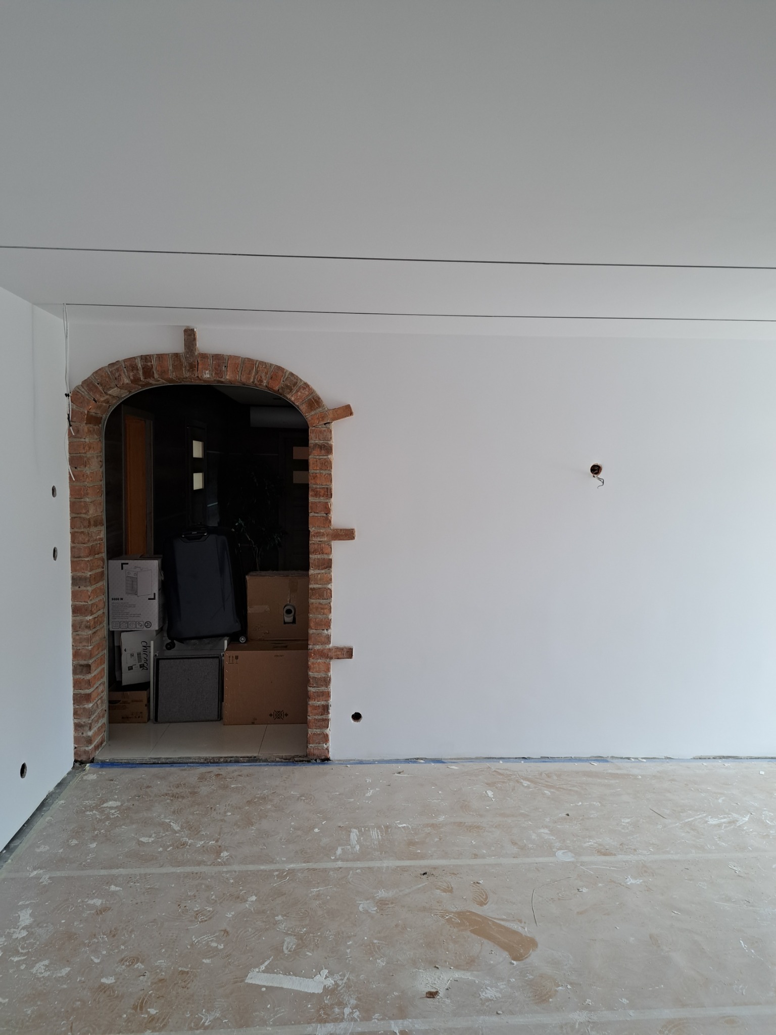 Wnętrze w trakcie prac wykończeniowych: surowe ściany pomalowane na biało, podłoga zabezpieczona folią, ceglany łuk wejścia do sąsiedniego pomieszczenia, w którym znajdują się kartony i walizka.