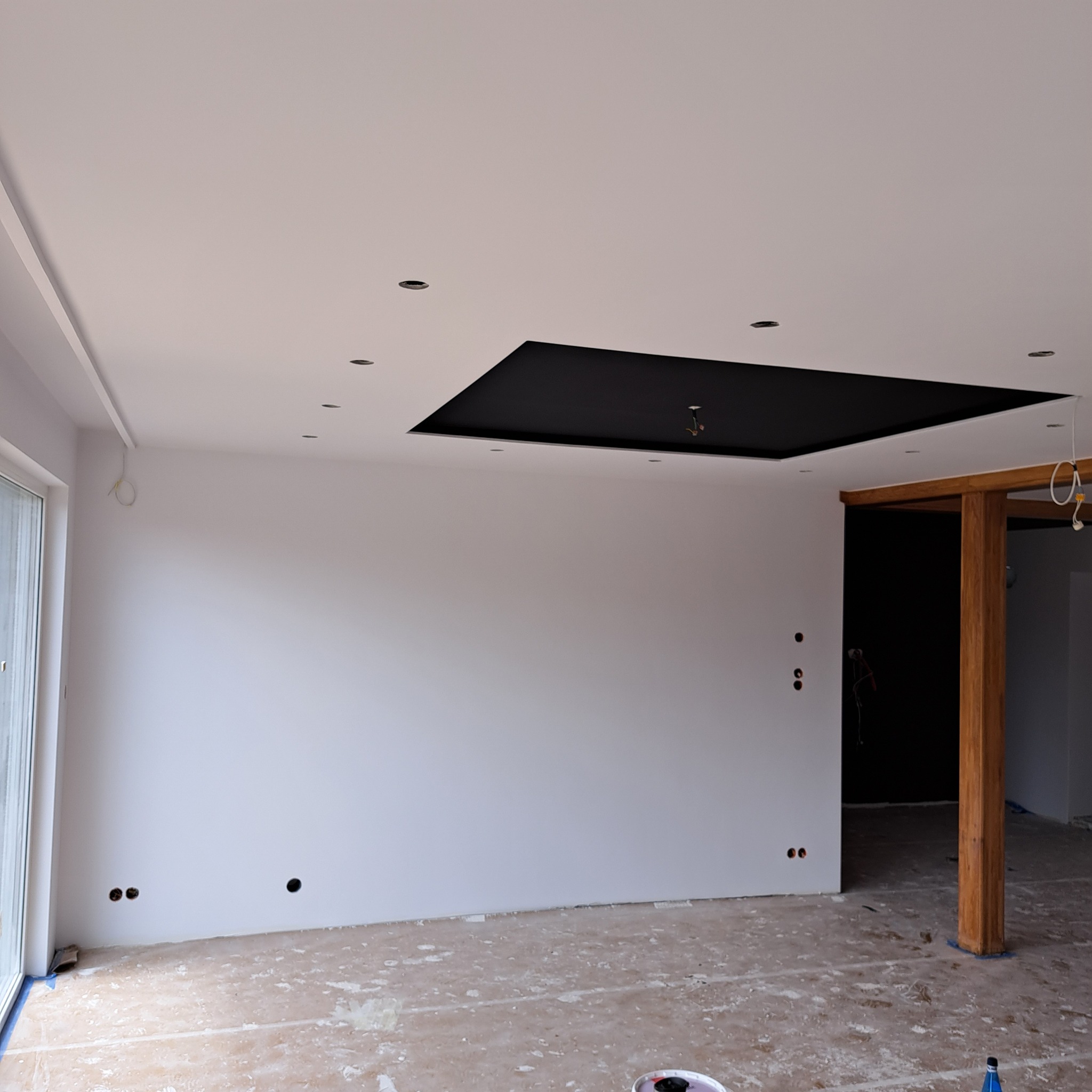 Wnętrze w trakcie malowania, widoczne świeżo pomalowane białe ściany, czarny fragment sufitu z oświetleniem punktowym, zabezpieczona podłoga i drewniany filar.