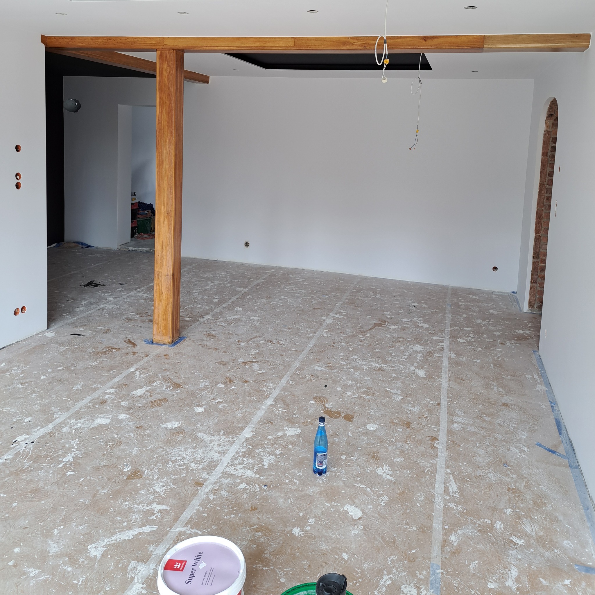 Wnętrze w trakcie malowania: białe ściany, drewniane belki stropowe, podłoga z widocznymi śladami farby, zabezpieczona taśmą malarską, butelka wody i puszka z farbą 'Super White' na podłodze.