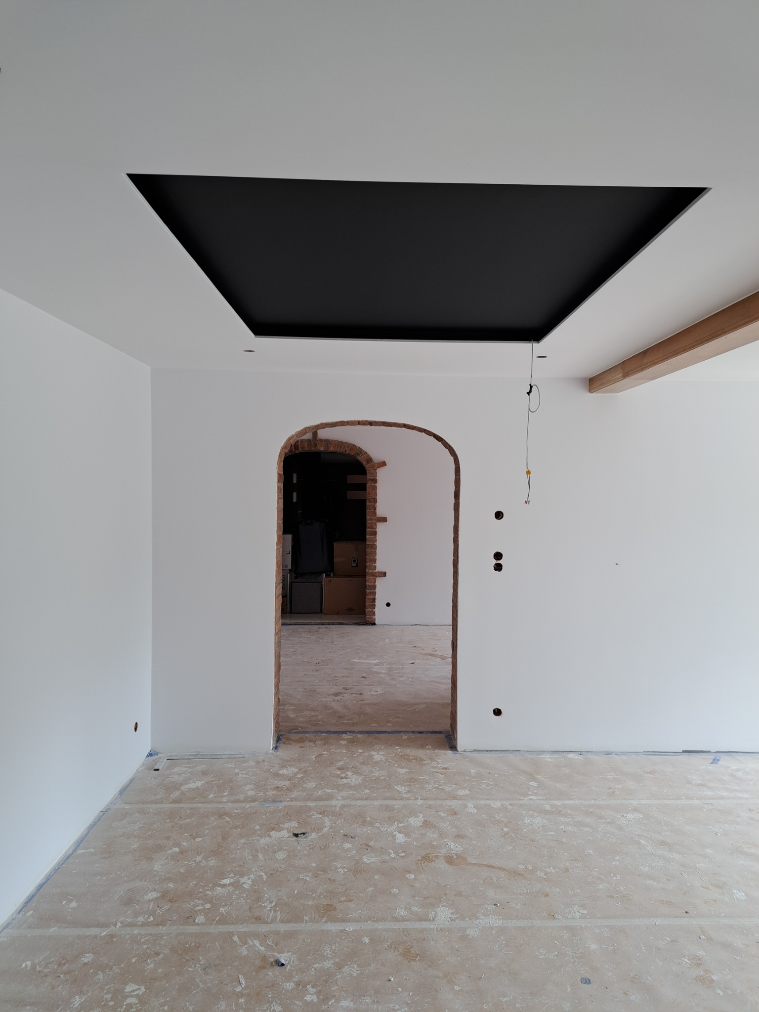 Wnętrze w trakcie prac wykończeniowych: białe ściany, czarny sufit podwieszany z oświetleniem punktowym, łukowe przejście z cegły, zabezpieczona podłoga, widoczne kable i puszki elektryczne.