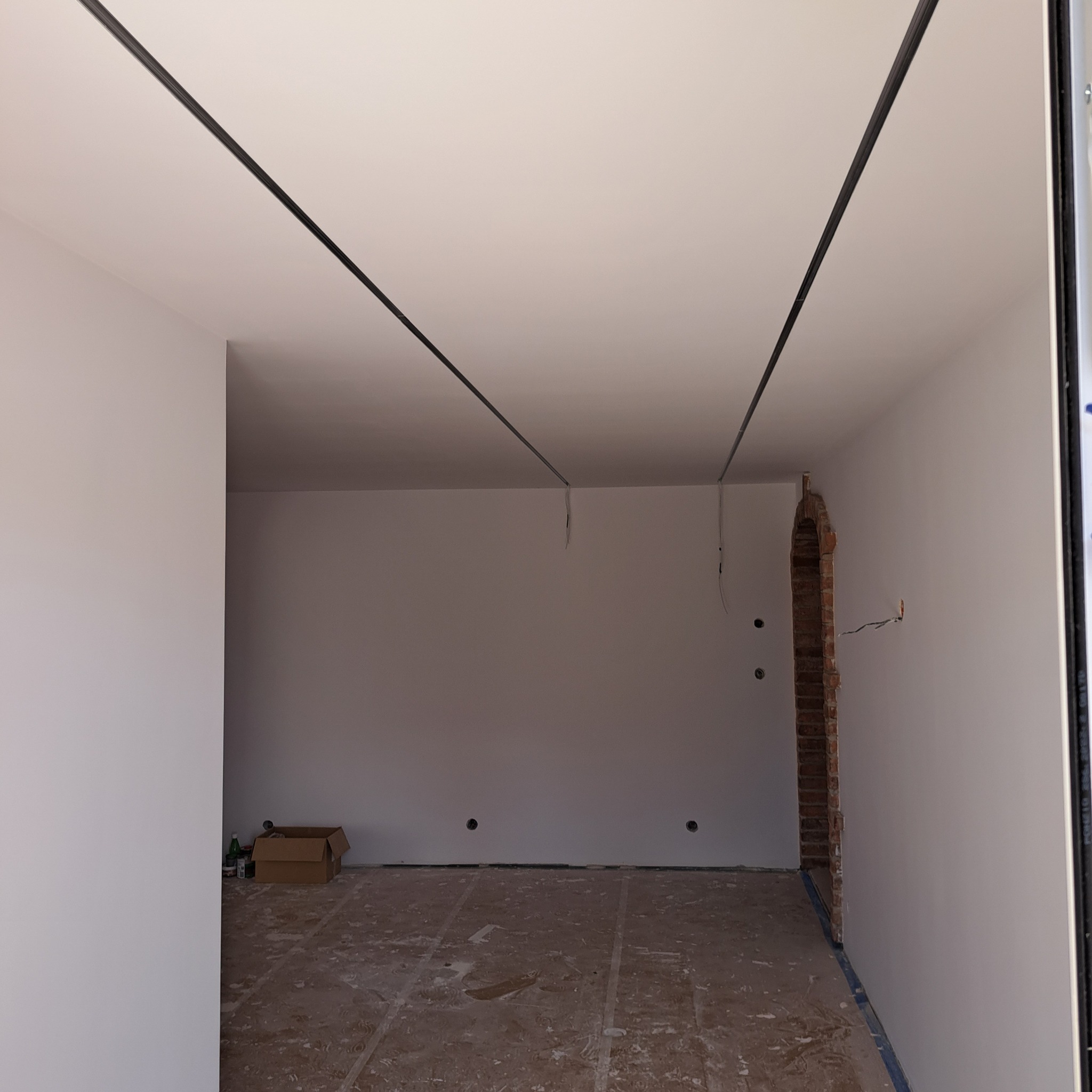 Wnętrze pokoju w trakcie remontu: białe ściany, sufit z czarnymi listwami, odsłonięty fragment ceglanego łuku, podłoga z resztkami zaprawy, karton i butelki w rogu.