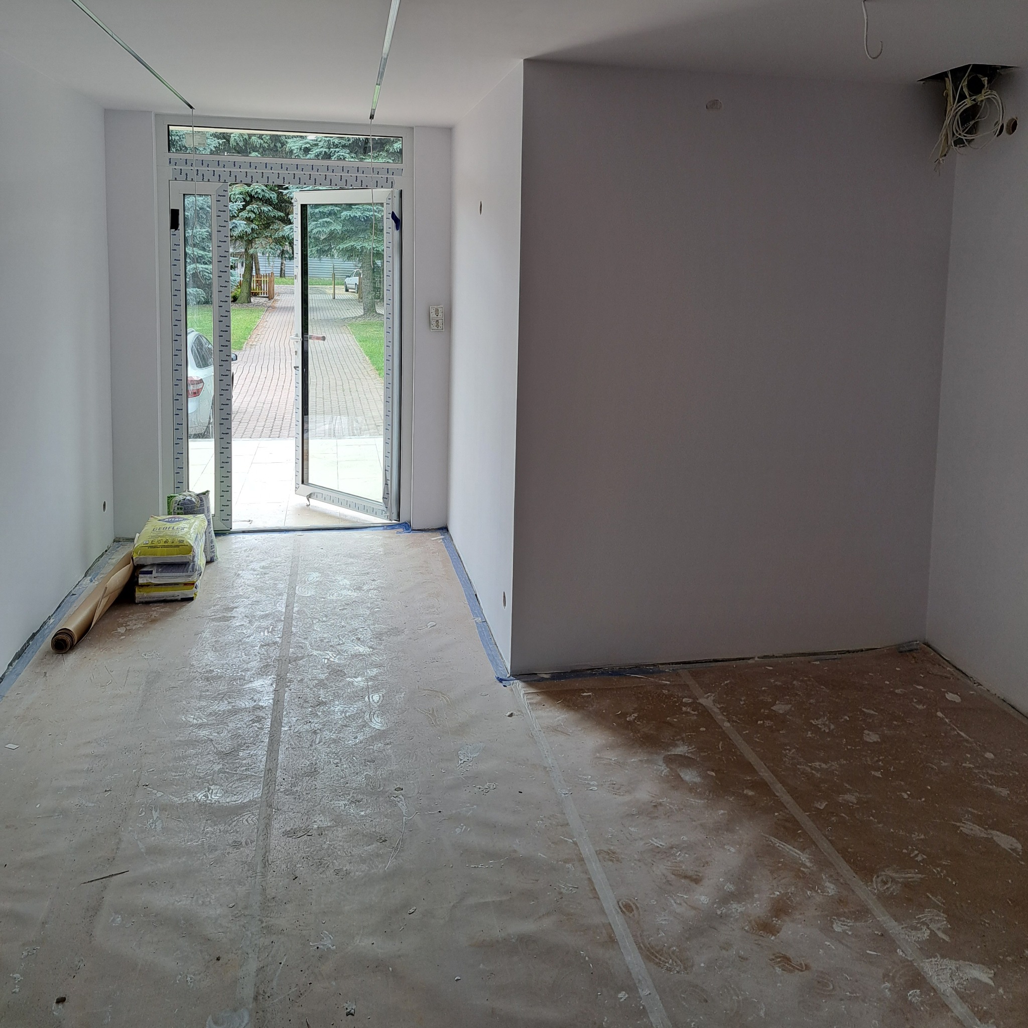 Przygotowane do remontu wnętrze z zabezpieczoną folią podłogą, workami z zaprawą przy otwartym oknie balkonowym i widocznymi przewodami elektrycznymi w rogu.