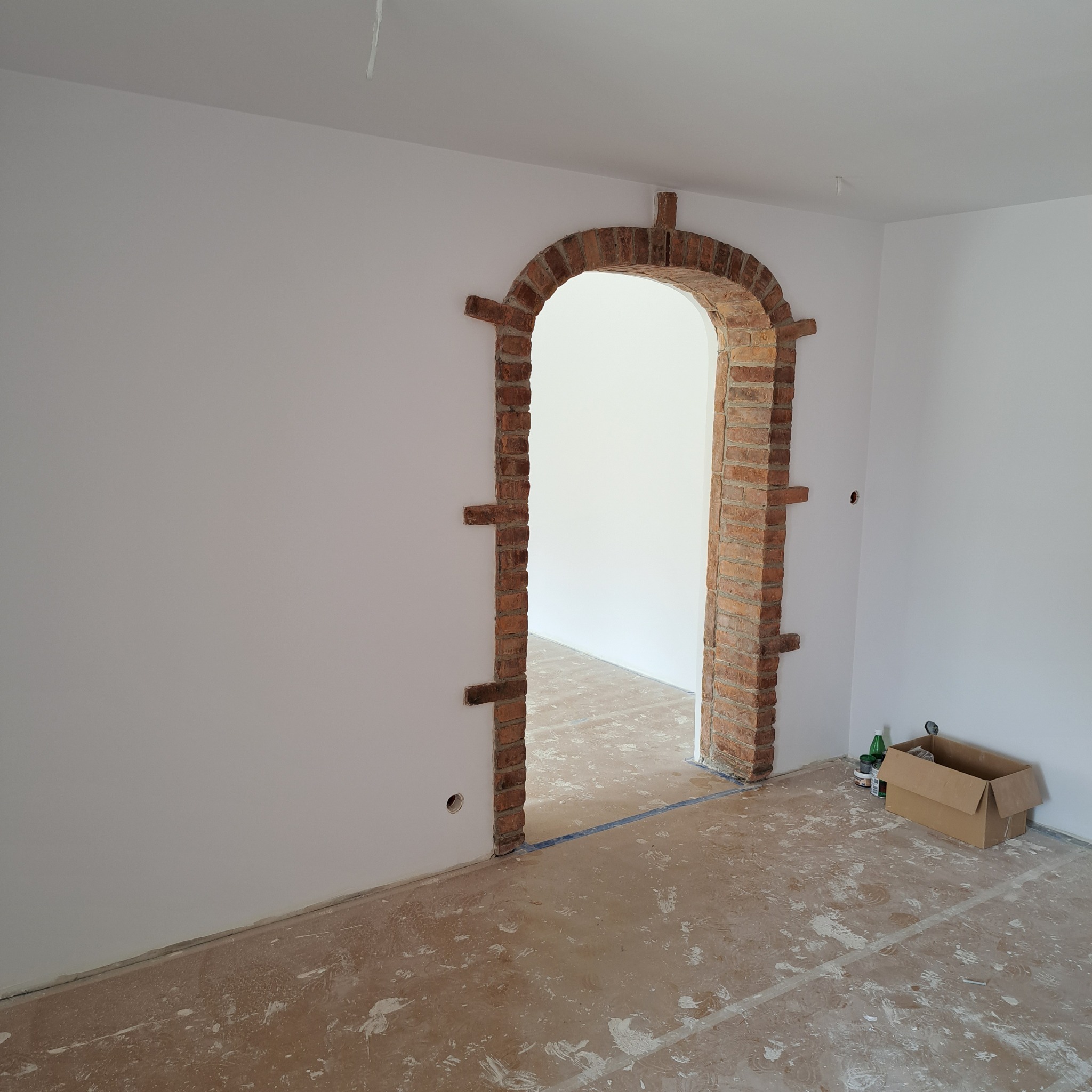 Wnętrze w trakcie prac: surowa ceglana rama łukowego przejścia między pokojami, świeżo pomalowane białe ściany, przygotowane podłoże pod posadzkę, karton i butelki po napojach w rogu.