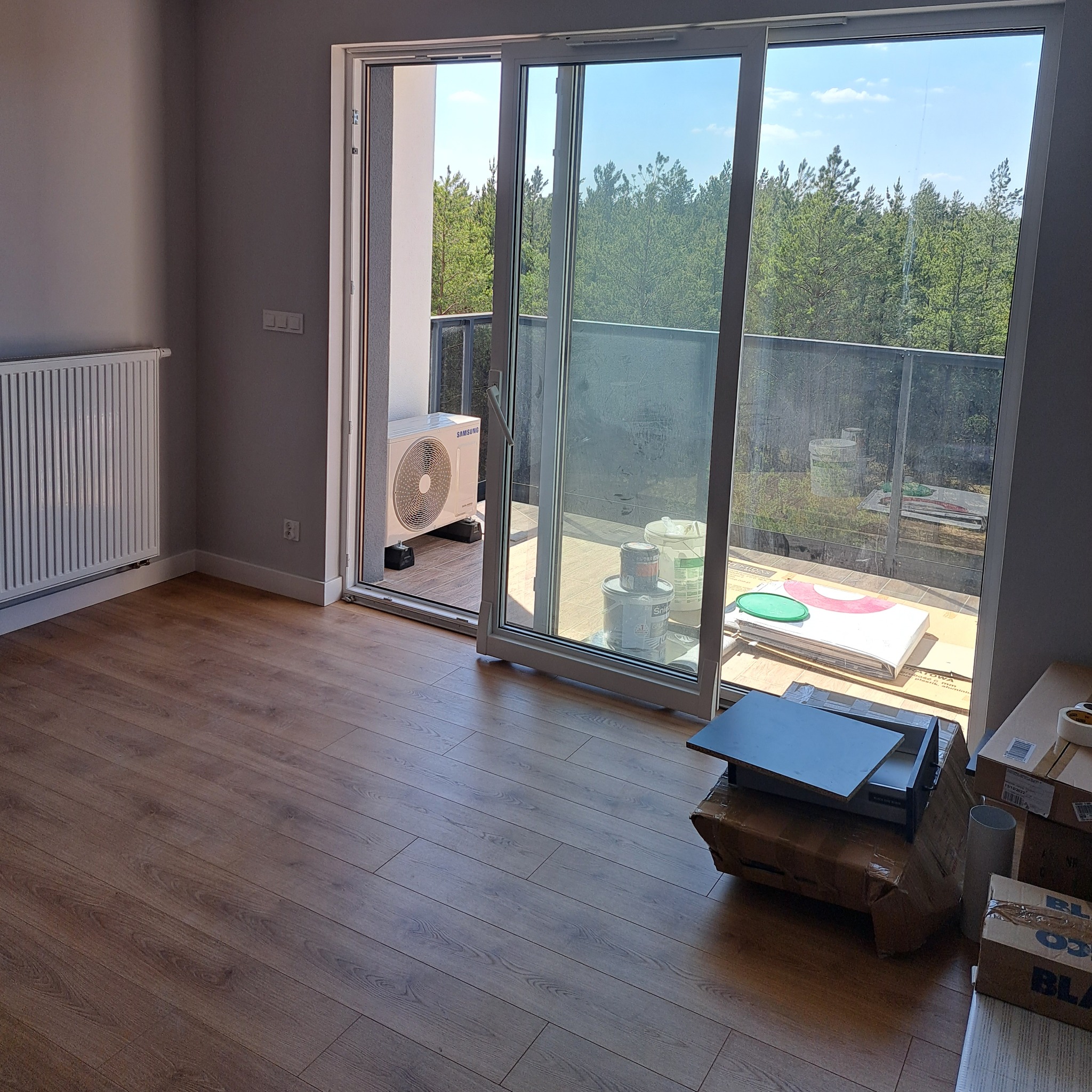 Pusty pokój z panelami podłogowymi w odcieniach brązu, grzejnik na ścianie, otwarte przesuwne drzwi balkonowe z widokiem na balkon, gdzie stoi klimatyzator Samsung, puszki z farbą i kartony...