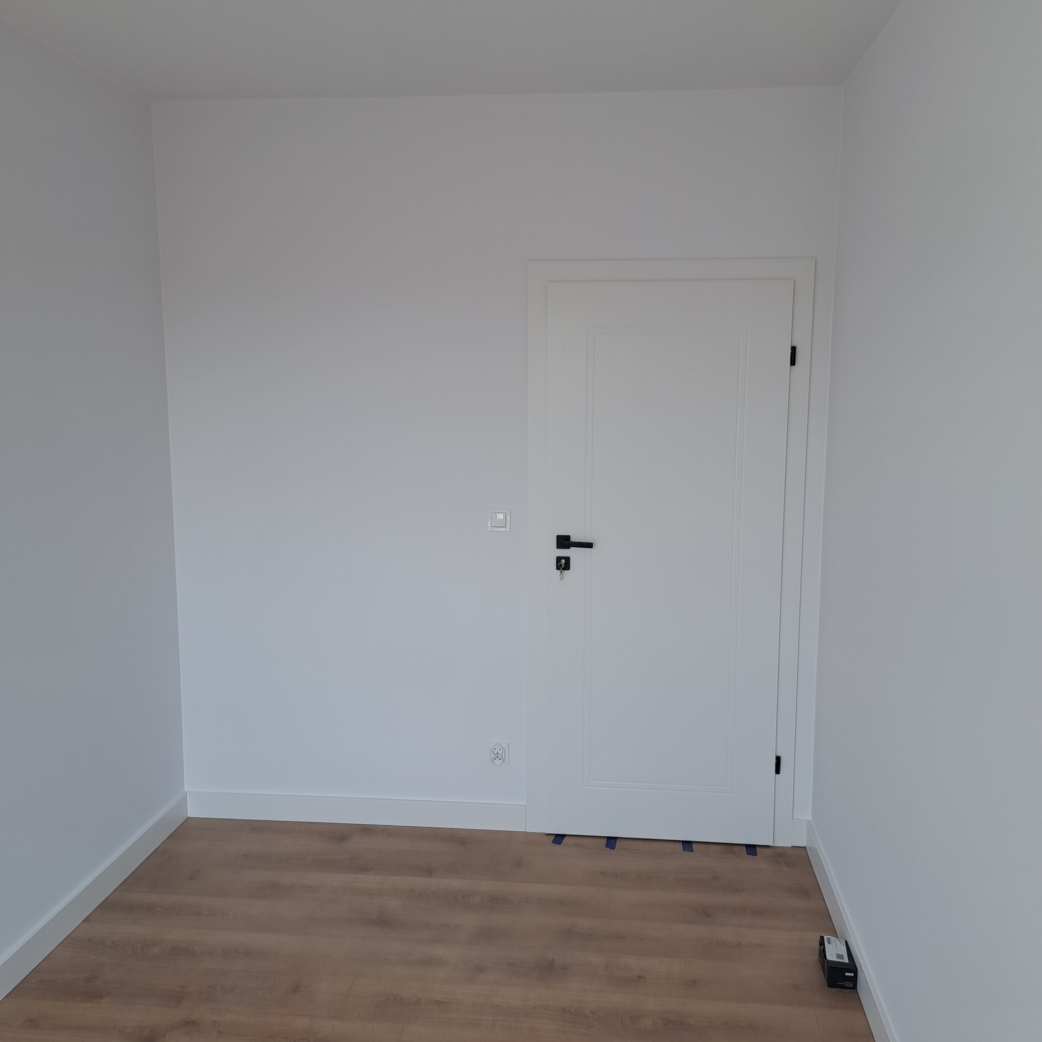 Pomalowane na biało wnętrze pokoju z panelami podłogowymi, białą listwą przypodłogową i nowymi białymi drzwiami z czarną klamką. Na ścianie widoczny włącznik światła i gniazdko. Pod drzwiami...