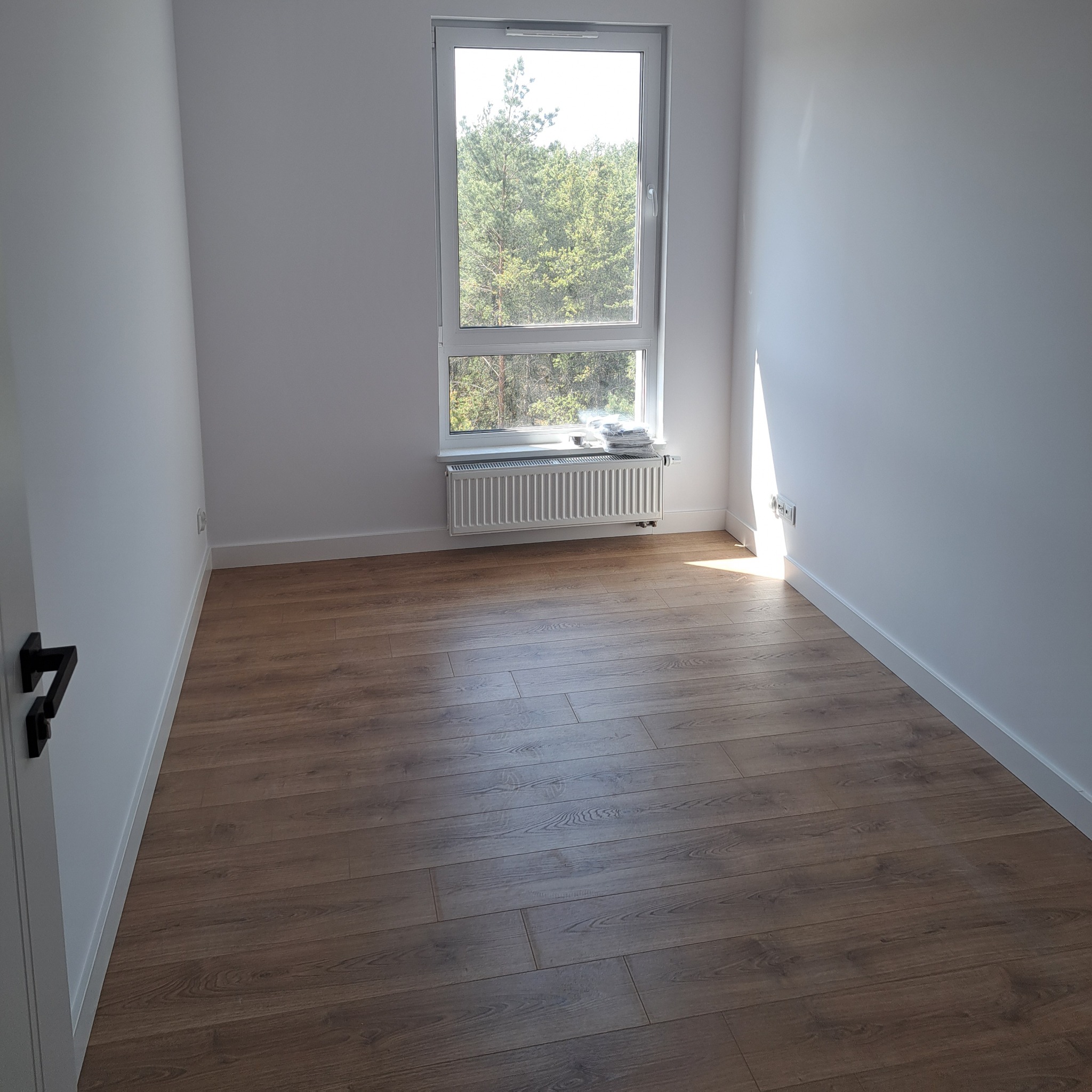 Pusty pokój po remoncie z panelami podłogowymi w odcieniach brązu, białymi ścianami, grzejnikiem pod oknem i widokiem na drzewa.