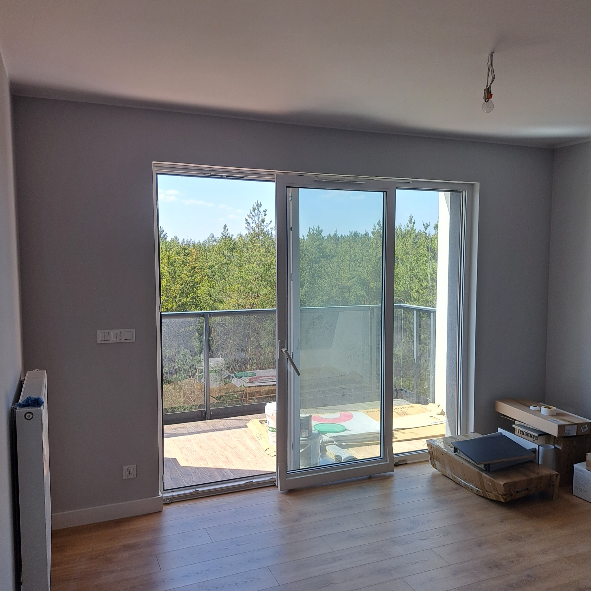 Pusty pokój z drewnianą podłogą, szarymi ścianami i otwartymi przesuwnymi drzwiami balkonowymi, przez które widać drzewa; widoczny grzejnik, gniazdka elektryczne, wisząca żarówka i kartony...