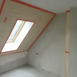 Poddasze w trakcie ocieplania z widocznym oknem dachowym, drewnianą boazerią zabezpieczoną taśmą malarską i częściowo wykończonymi ścianami.