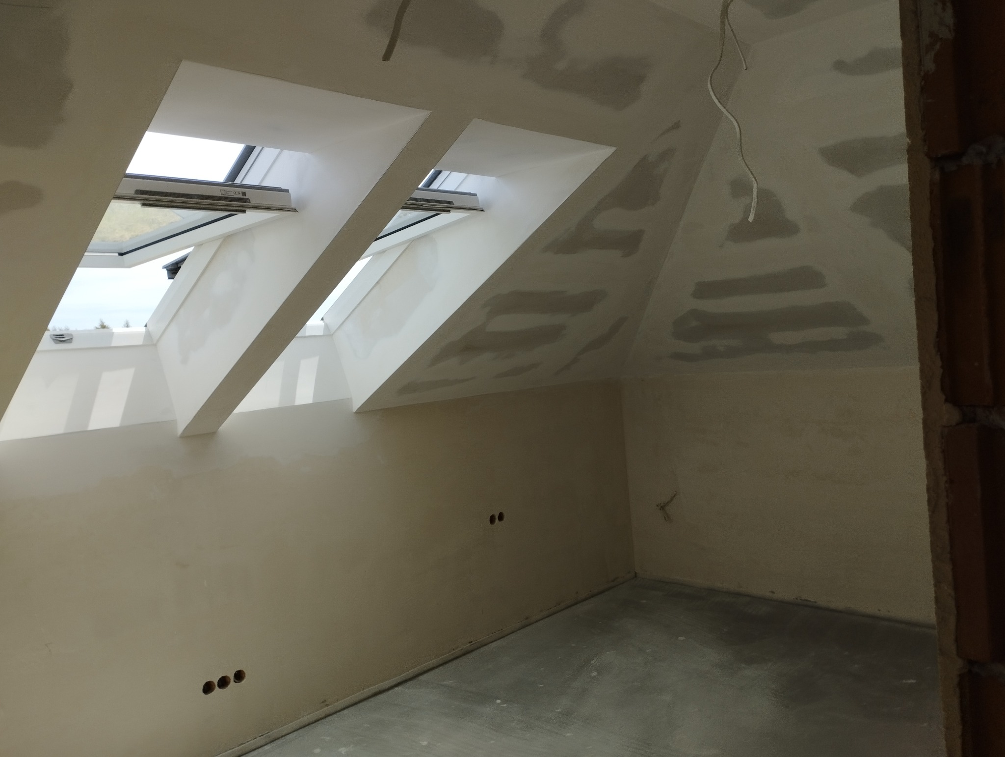 Poddasze w trakcie prac wykończeniowych, widoczne surowe ściany po nałożeniu gładzi, okna dachowe i przygotowane otwory na gniazdka elektryczne.