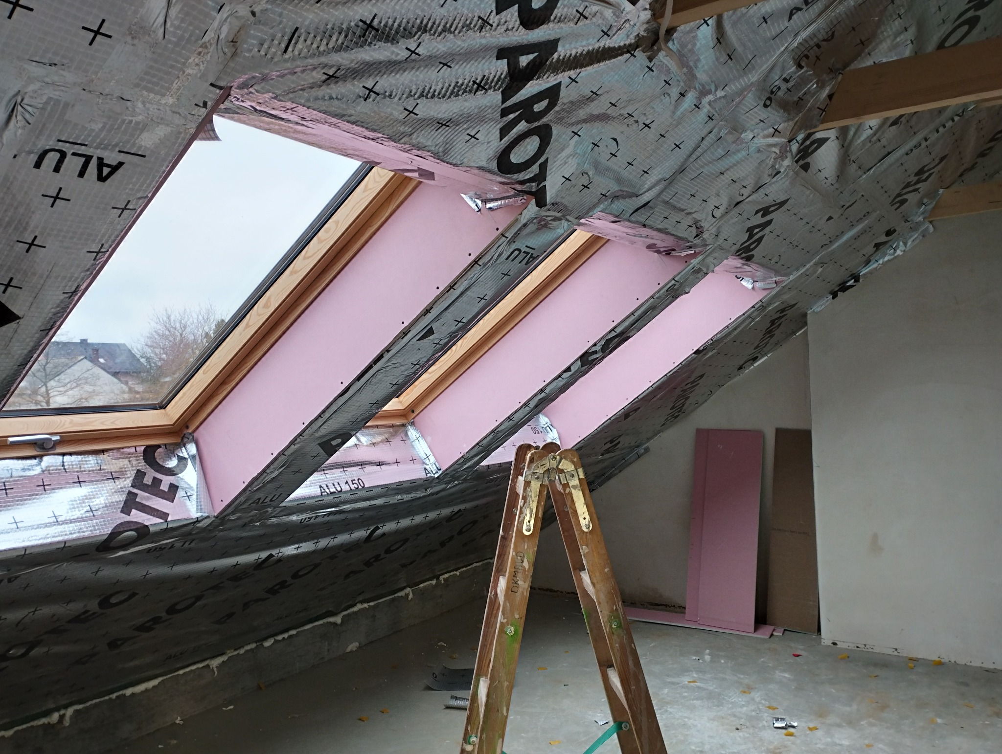 Montaż izolacji poddasza z widocznymi oknami dachowymi, folią paroizolacyjną i różową wełną mineralną, drabina w pomieszczeniu.