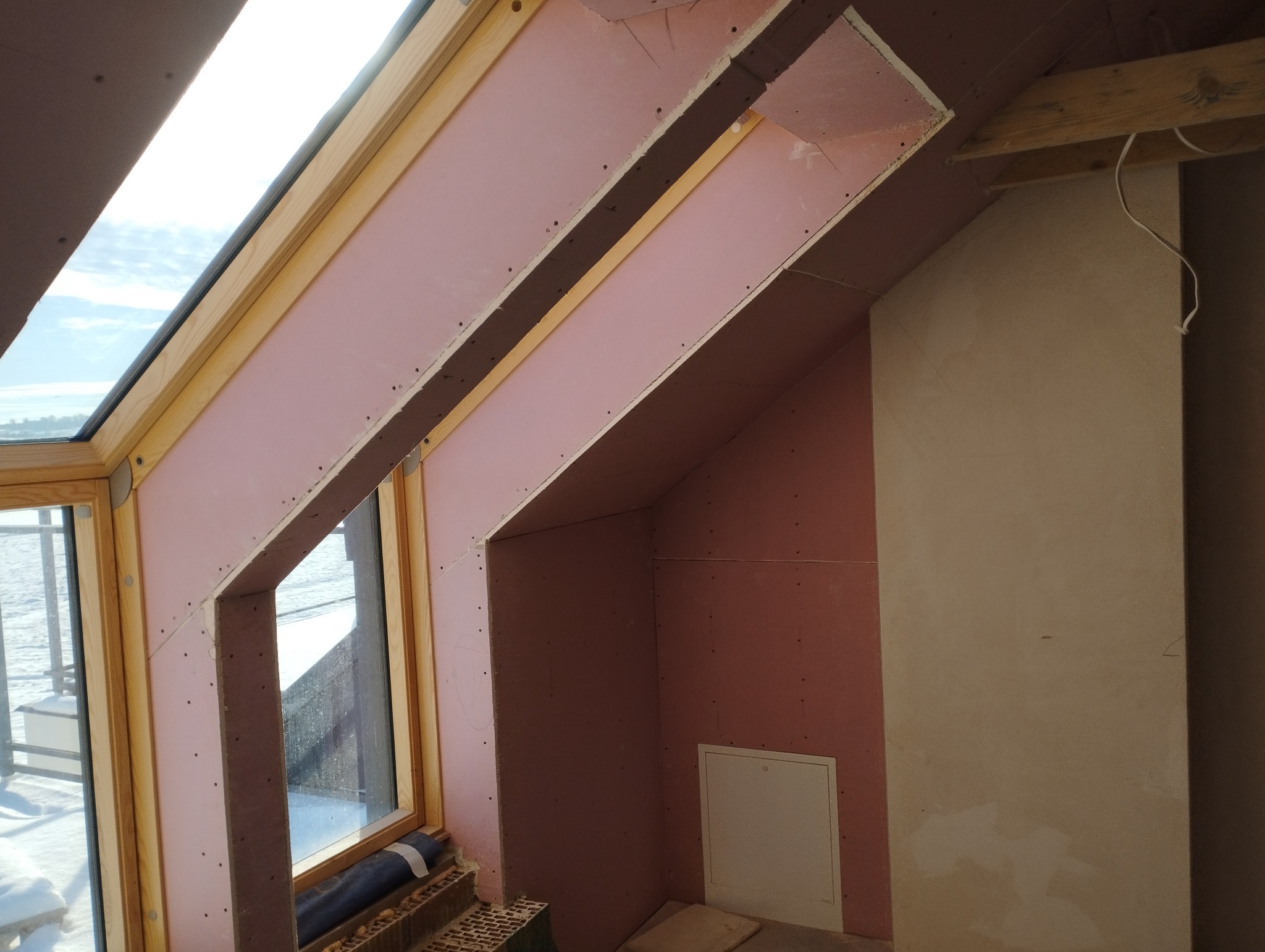 Poddasze z oknem dachowym w trakcie montażu płyt kartonowo-gipsowych; widoczne drewniane ramy okienne, fragment ocieplenia oraz ściana przygotowana pod gładzie.