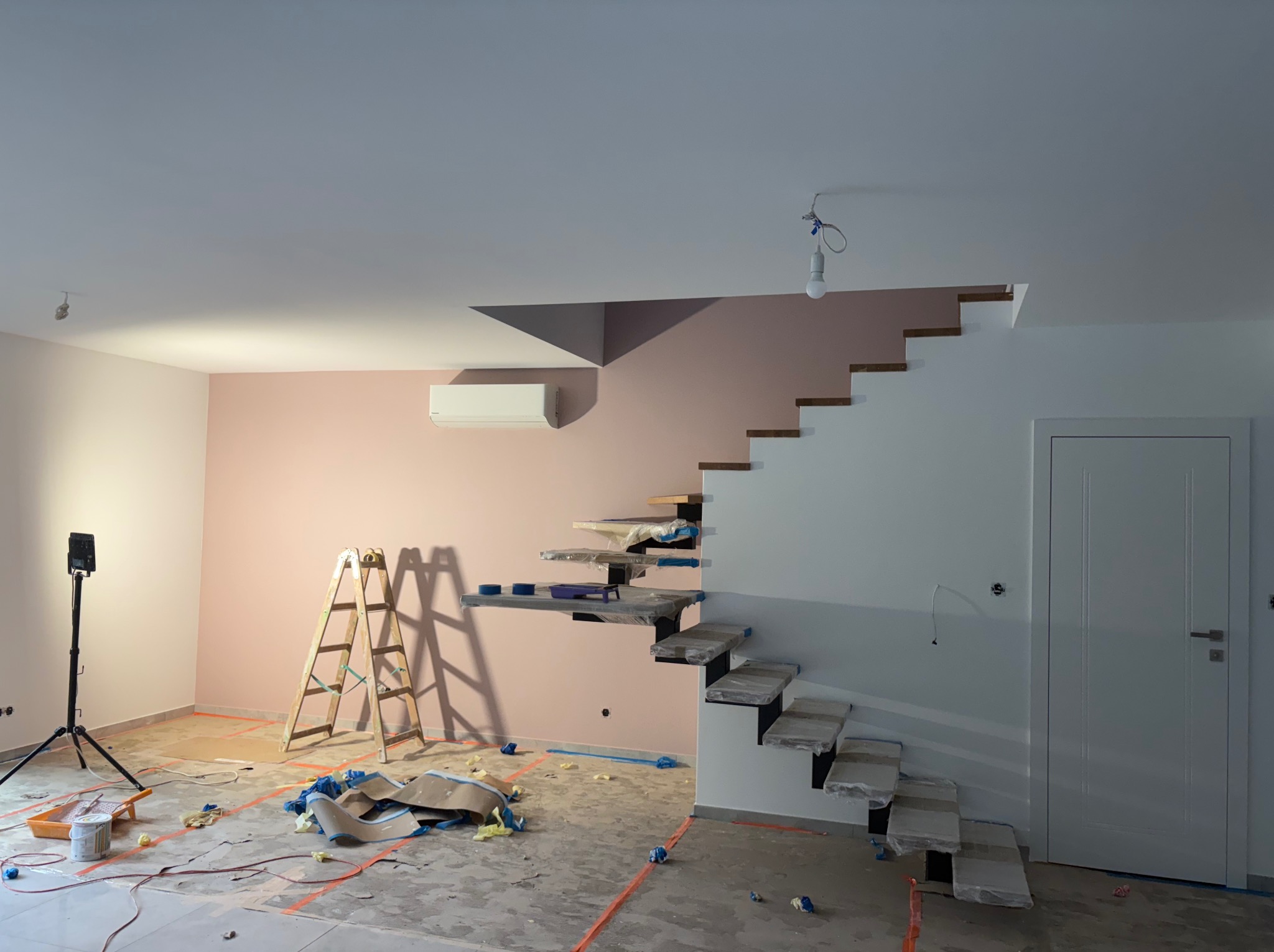 Wnętrze w trakcie malowania: ściana w odcieniach różu, schody zabezpieczone folią, drabina i lampa na statywie. Widoczne ślady farby na podłodze.