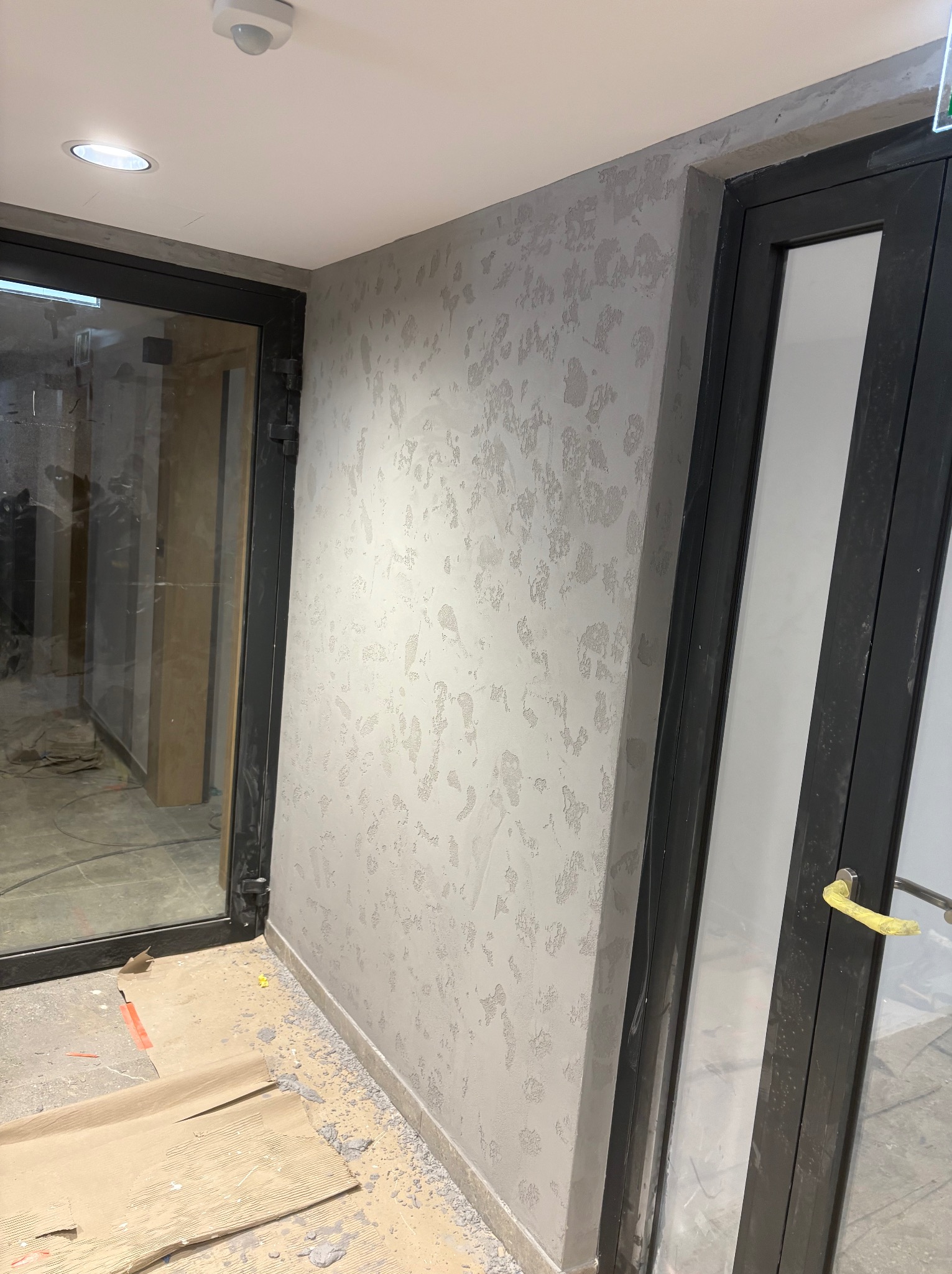 Ściana z dekoracyjną, szarą strukturą obok czarnej ramy drzwi. Na podłodze karton zabezpieczający. Widoczny fragment sufitu z oświetleniem.