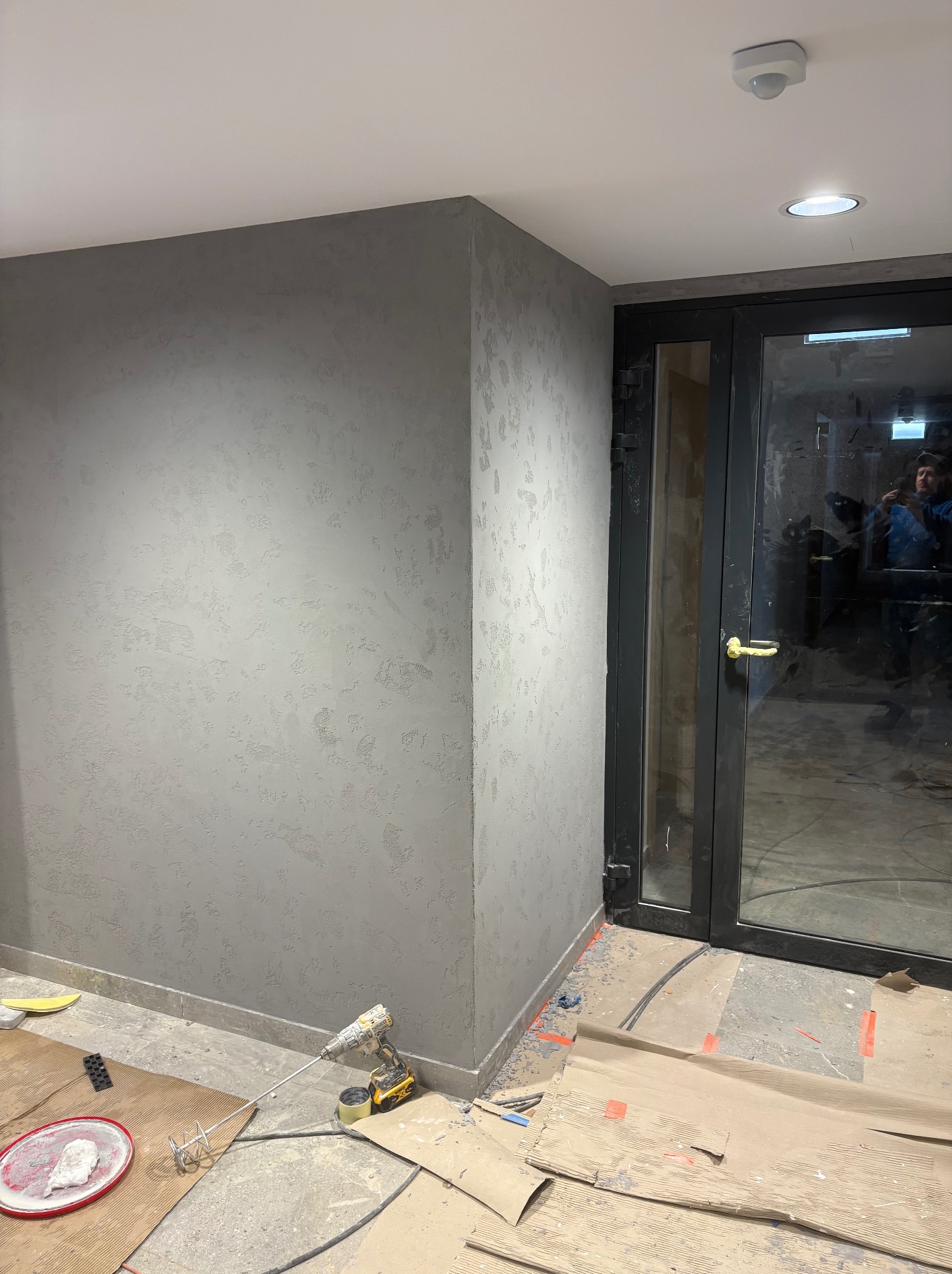 Ściana z szarą, dekoracyjną strukturą tynku w rogu pomieszczenia, obok czarnych drzwi. Na podłodze narzędzia i zabezpieczenia malarskie.