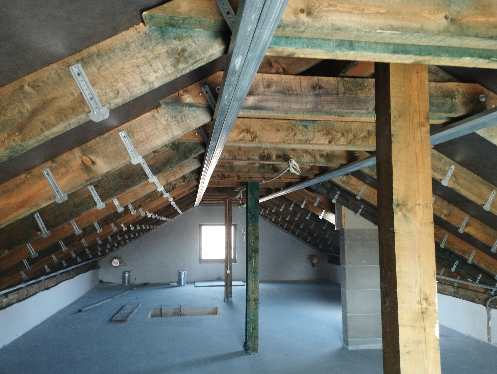 Poddasze w trakcie adaptacji: drewniana konstrukcja dachu z zamocowanymi metalowymi wspornikami, widoczne słupy nośne i profile metalowe, przygotowane do dalszych prac wykończeniowych, w tle okno.