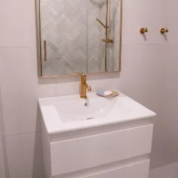 Nowoczesna łazienka z białą szafką pod umywalką, złotą baterią i lustrem z widoczną kabiną prysznicową wyłożoną biało-szarą mozaiką w jodełkę.