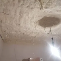 Poddasze zaizolowane pianką, widoczne wystające kable elektryczne i niedokończone wykończenie, oświetlone mocnym światłem z dołu.