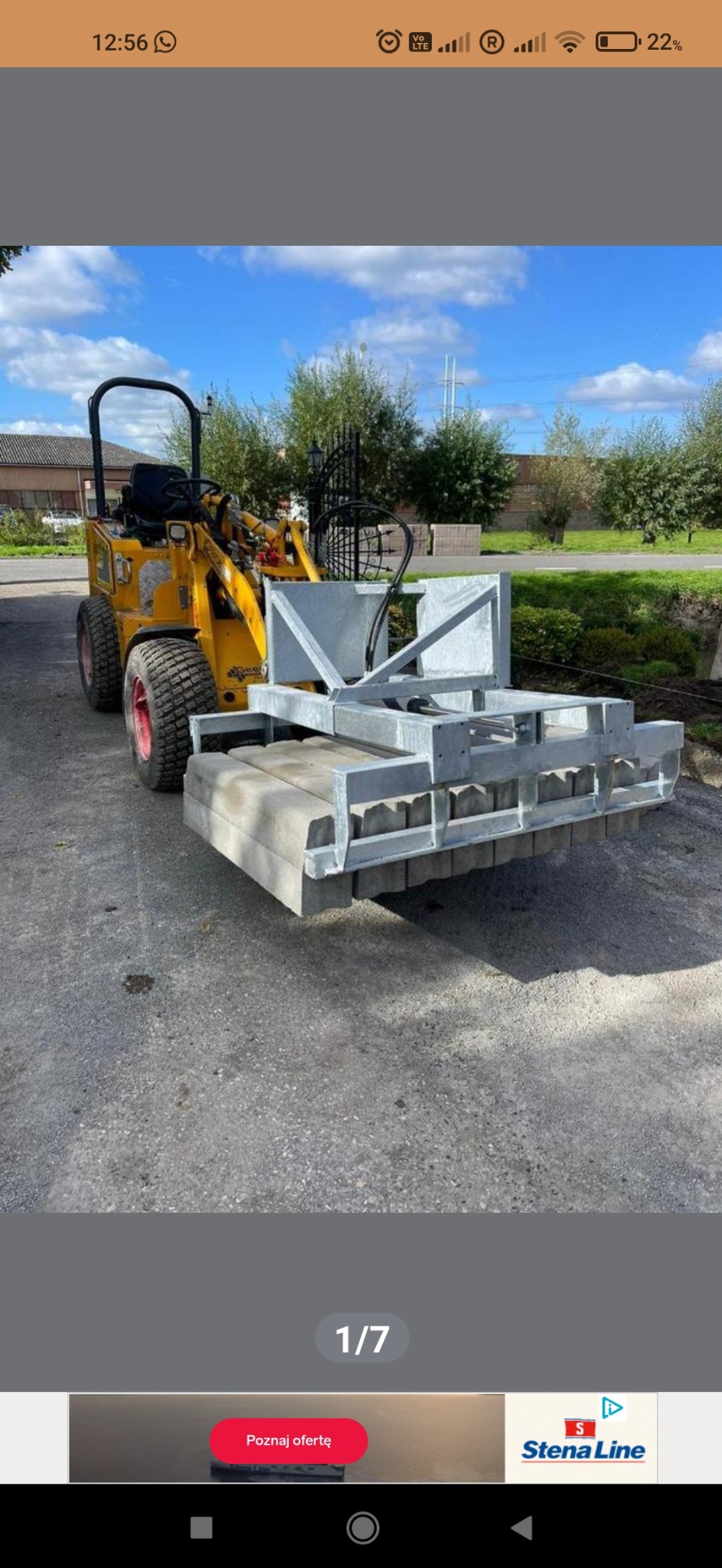 Żółta miniładowarka z zamontowanym osprzętem do układania obrzeży betonowych na utwardzonej powierzchni, widok z boku, w tle zabudowa jednorodzinna i zieleń.