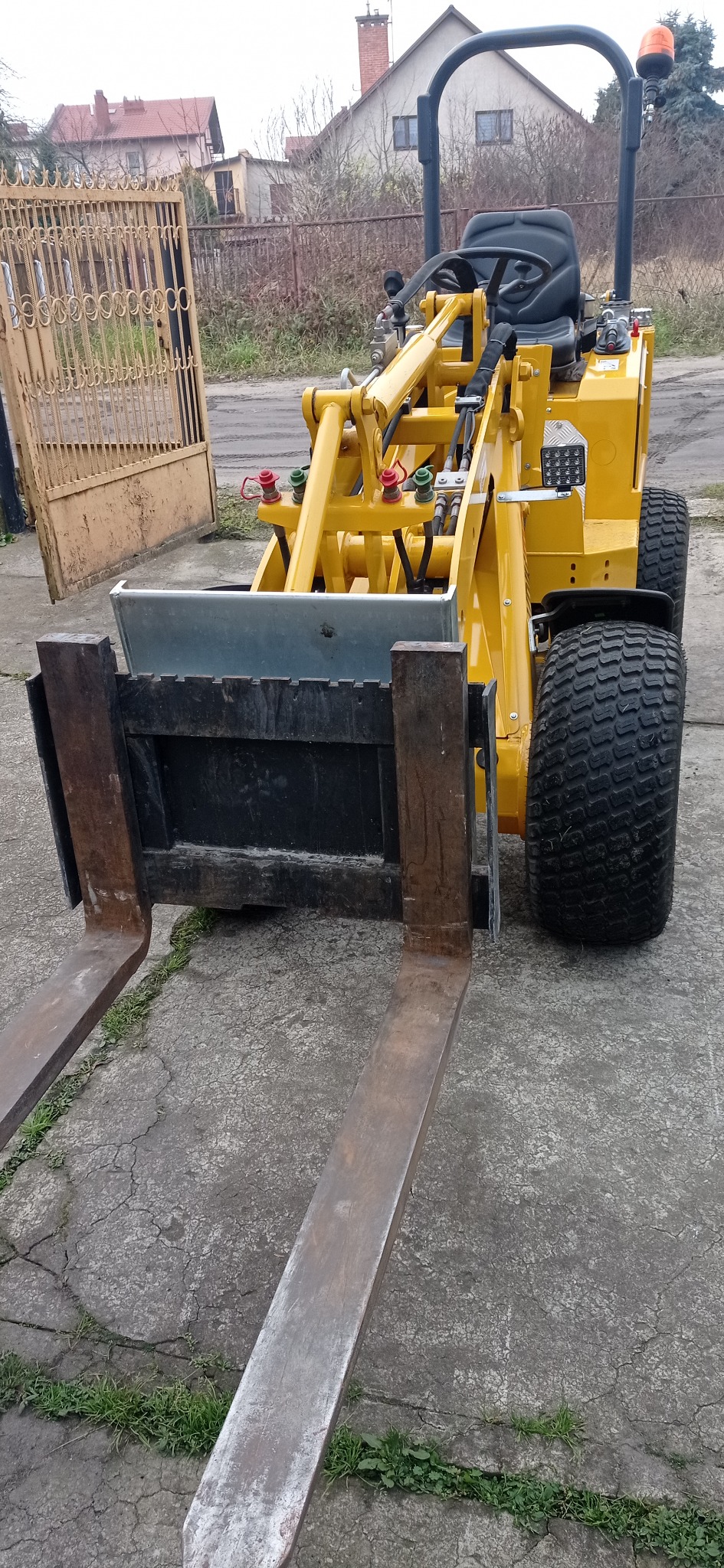 Żółta mini ładowarka teleskopowa z widłami, zaparkowana na betonowym podłożu przed otwartą, metalową bramą i budynkami w tle.