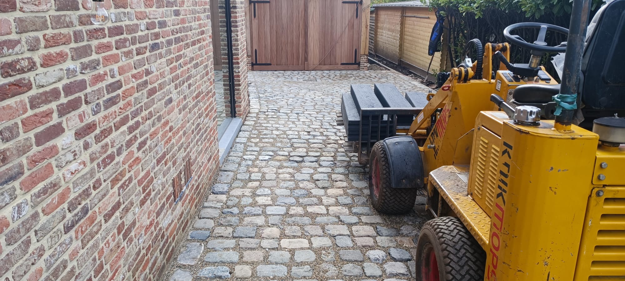 Ułożona kostka granitowa na podjeździe, widoczna maszyna budowlana Knikmops przewożąca elementy, ceglana ściana budynku w tle.