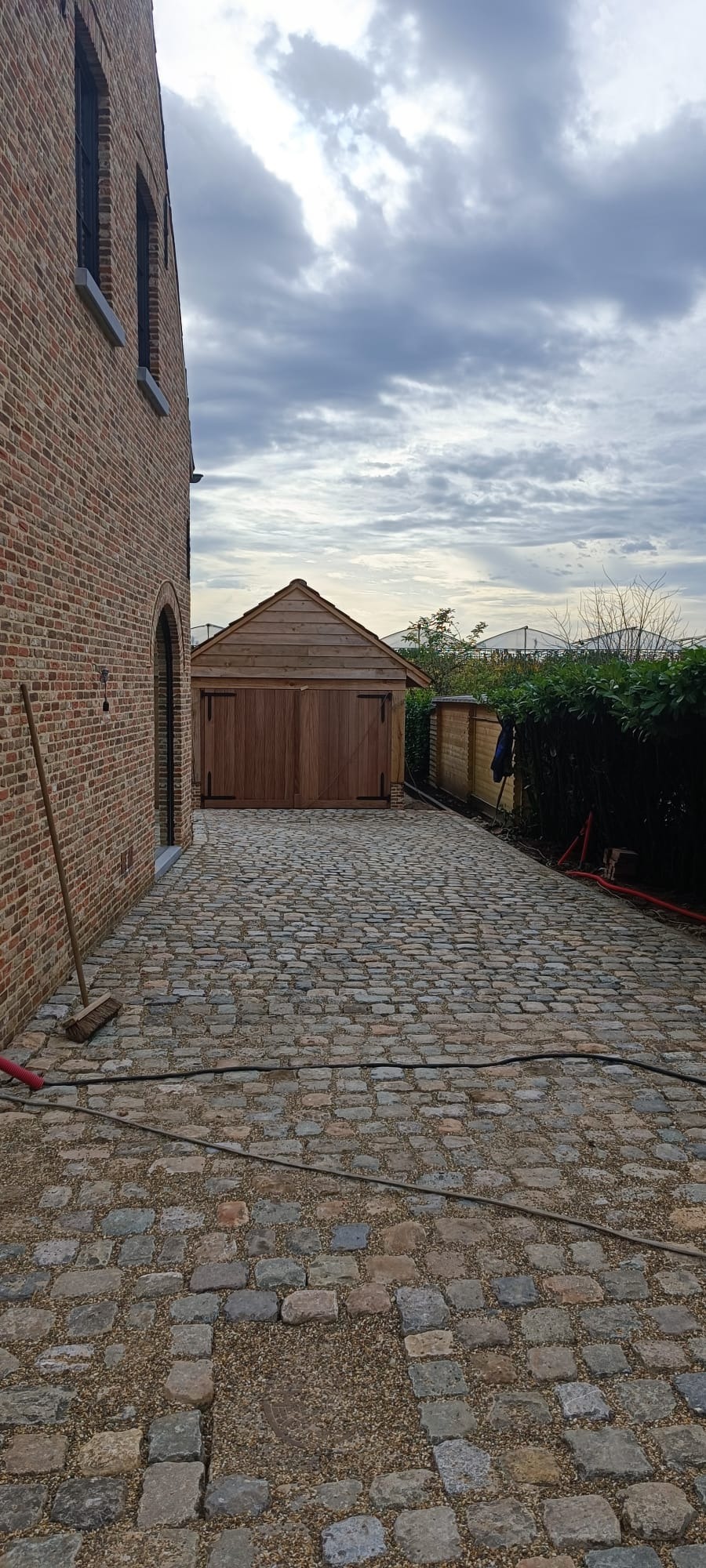 Ułożona kostka granitowa na podjeździe do drewnianego garażu, widok z perspektywy chodnika, obok ceglany budynek i żywopłot.