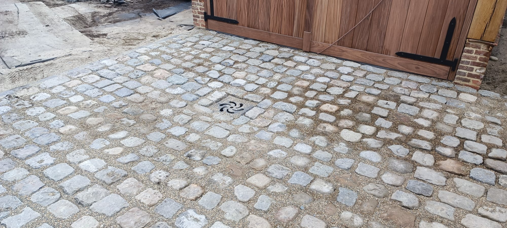 Ułożona nawierzchnia z kostki granitowej w odcieniach szarości, z widocznym odpływem liniowym i drewnianą bramą w tle.