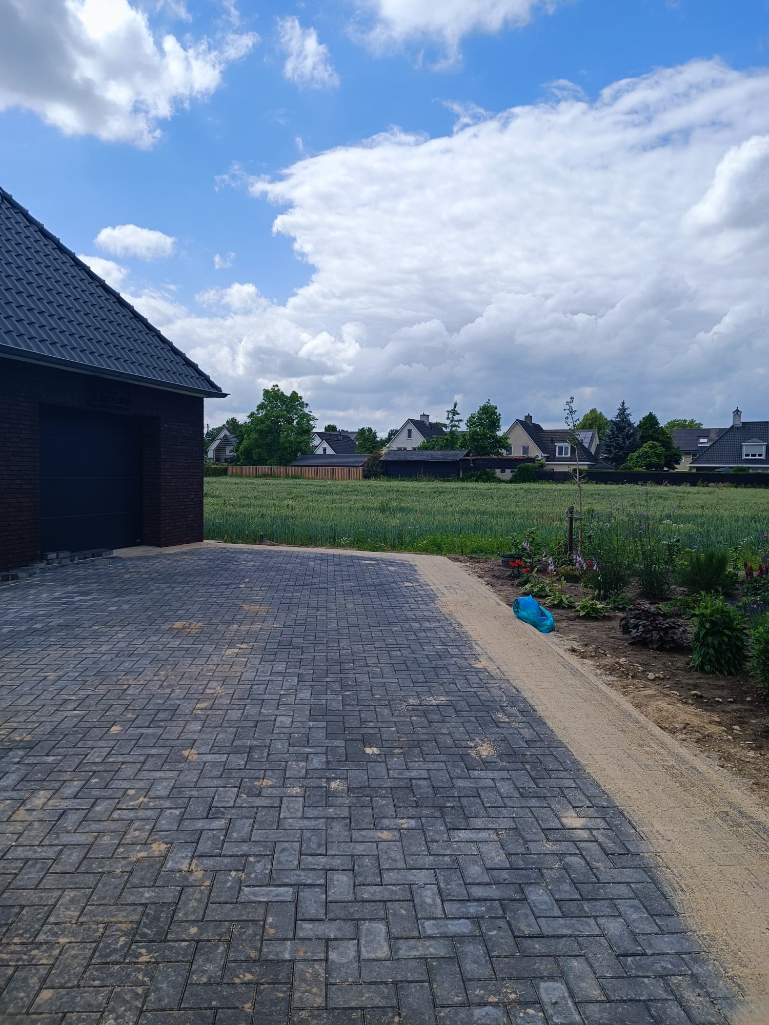 Świeżo ułożona, grafitowa kostka brukowa na podjeździe, z piaskową obwódką i widokiem na zielone pole oraz domy w tle, pod błękitnym niebem z obłokami.