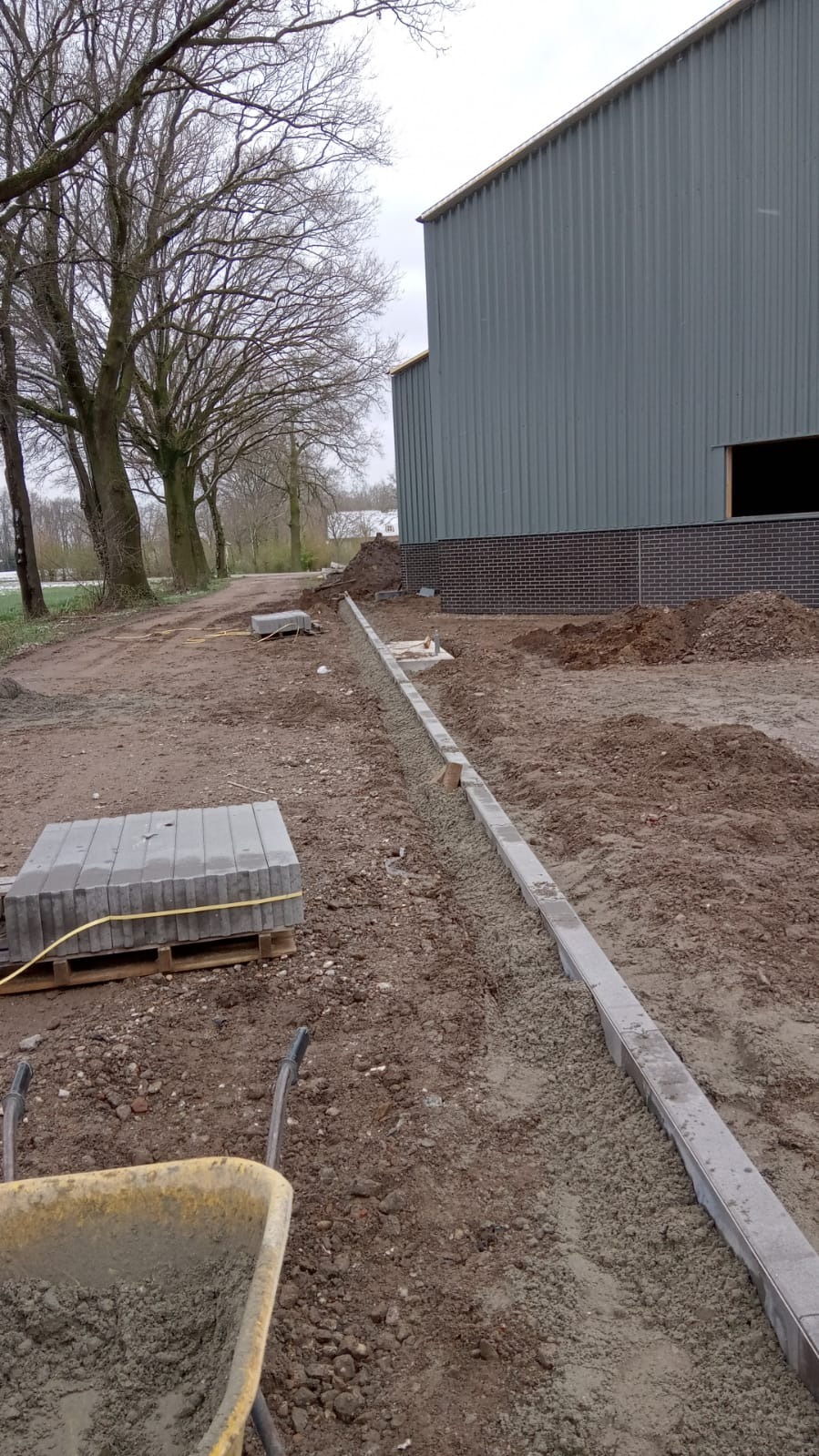 Przygotowanie podłoża pod ułożenie kostki: widoczny wykop z podsypką cementowo-piaskową, częściowo ułożone obrzeża betonowe, paleta z kostką brukową w tle, oraz taczka z zaprawą, teren budowy przy...