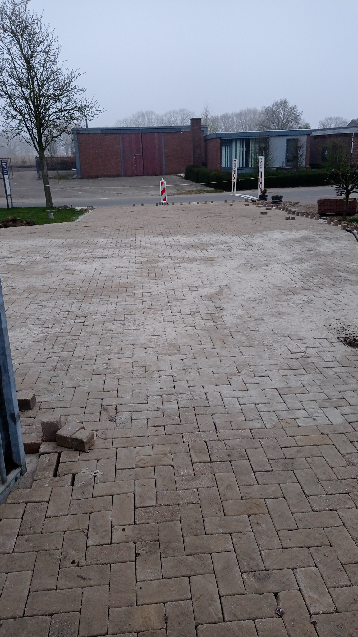 Układanie beżowej kostki brukowej w jodełkę na dużej powierzchni, widoczne niedokończone fragmenty i przygotowane kostki, w tle budynek gospodarczy i słupki ostrzegawcze.