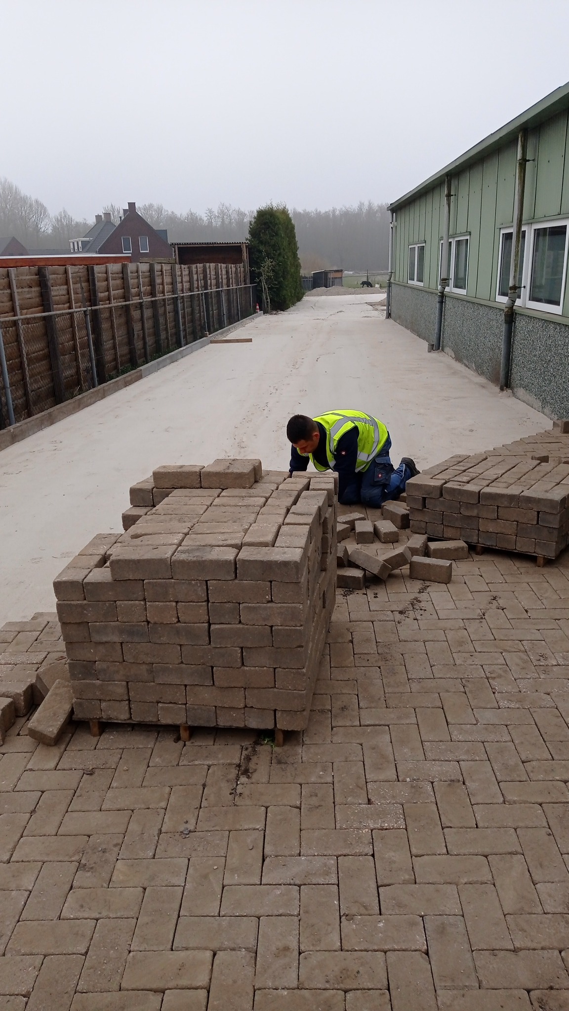 Pracownik w żółtej kamizelce układa kostkę brukową na posesji w Radomiu, widoczne palety z kostką i częściowo ułożona nawierzchnia.