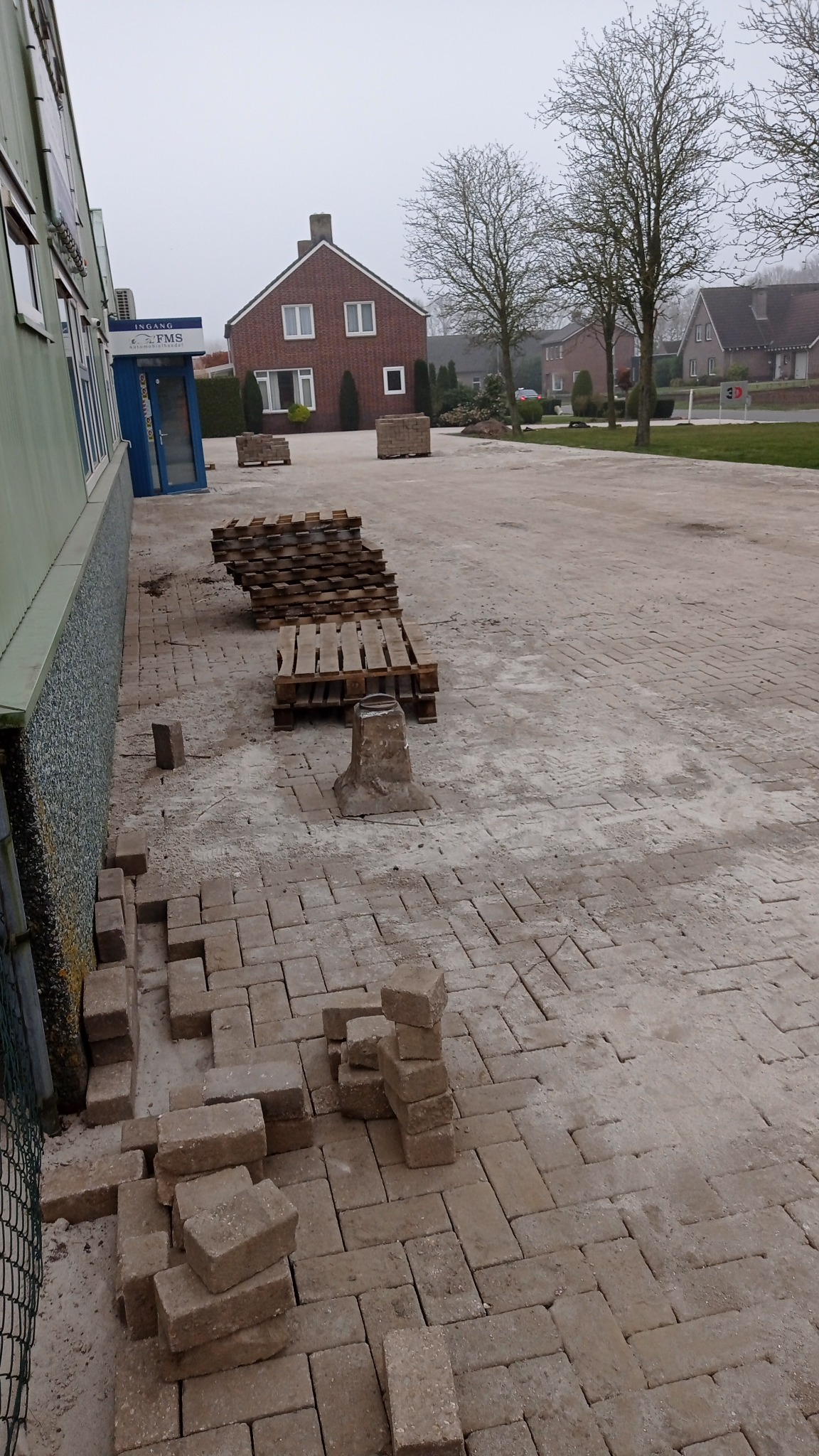 Ułożona część nawierzchni z kostki brukowej w jodełkę, palety z kostką w tle, budynek z zieloną elewacją po lewej, ceglany budynek mieszkalny w oddali, pochmurne niebo.