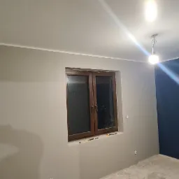 Pokój po malowaniu, ściany w odcieniach szarości i granatu, widoczne okno z drewnianą ramą, surowa podłoga przed wykończeniem.