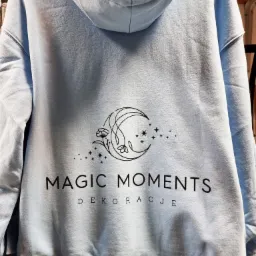 Jasnoniebieska bluza z kapturem, na plecach czarny nadruk z motywem księżyca, kwiatów i gwiazd, oraz napisy 'MAGIC MOMENTS DEKORACJE'.