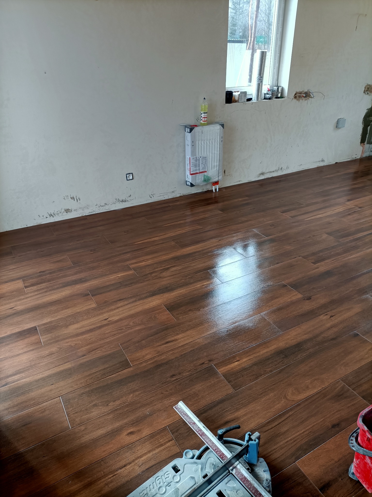 Ułożona podłoga z płytek imitujących drewno w nowym pomieszczeniu, widoczny sprzęt do cięcia płytek w rogu, ściany w trakcie przygotowania.