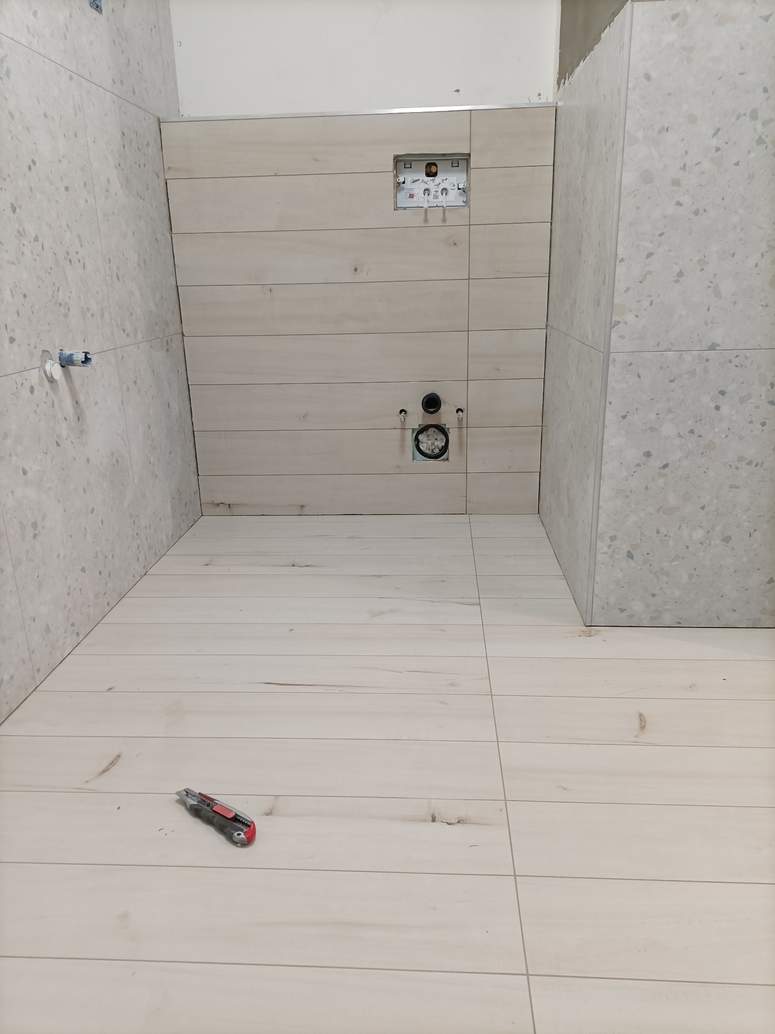 Łazienka w trakcie prac: ułożone jasne płytki imitujące drewno na podłodze i ścianie, widoczne otwory na instalacje sanitarne, nóż tapicerski leżący na podłodze.