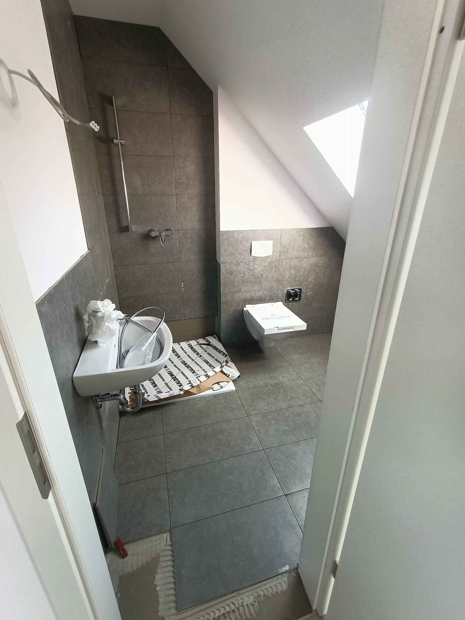 Remont łazienki w trakcie: szare płytki na ścianach i podłodze, biała umywalka z baterią i toaleta Villeroy & Boch jeszcze w opakowaniu, widok przez otwarte drzwi.