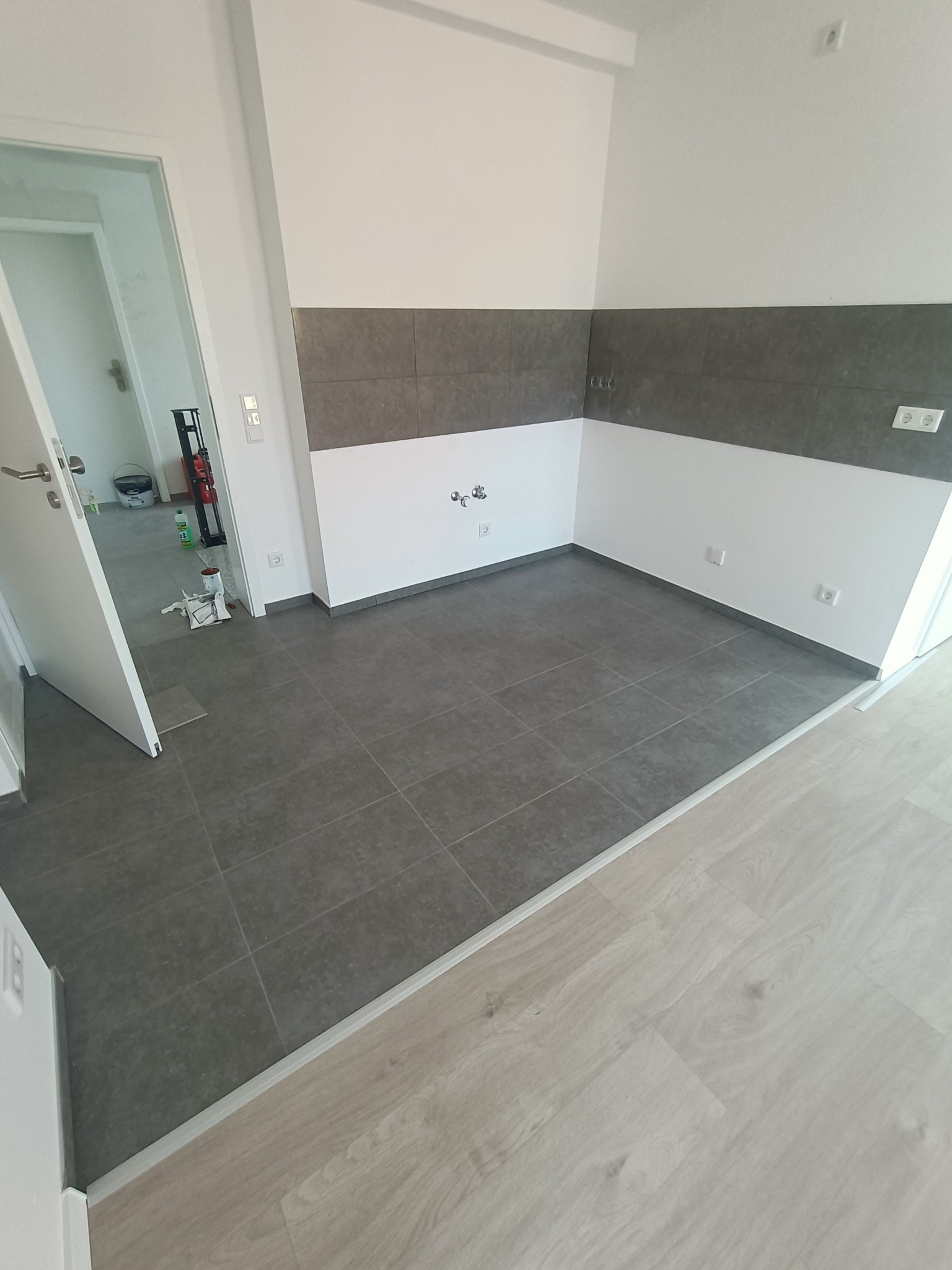 Ukończona podłoga z ciemnoszarych płytek w pomieszczeniu z białymi ścianami i szarym pasem płytek, widoczne gniazdka elektryczne i przejście na drewnianą podłogę.