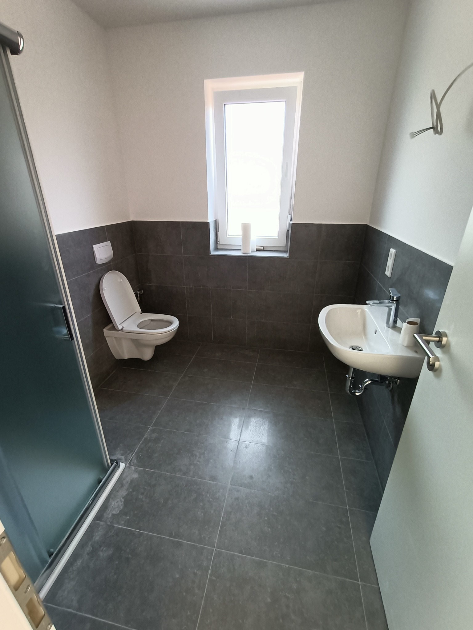 Nowoczesna łazienka z szarymi płytkami, białymi ścianami i armaturą, widok na umywalkę, toaletę i kabinę prysznicową z przeszkleniem.