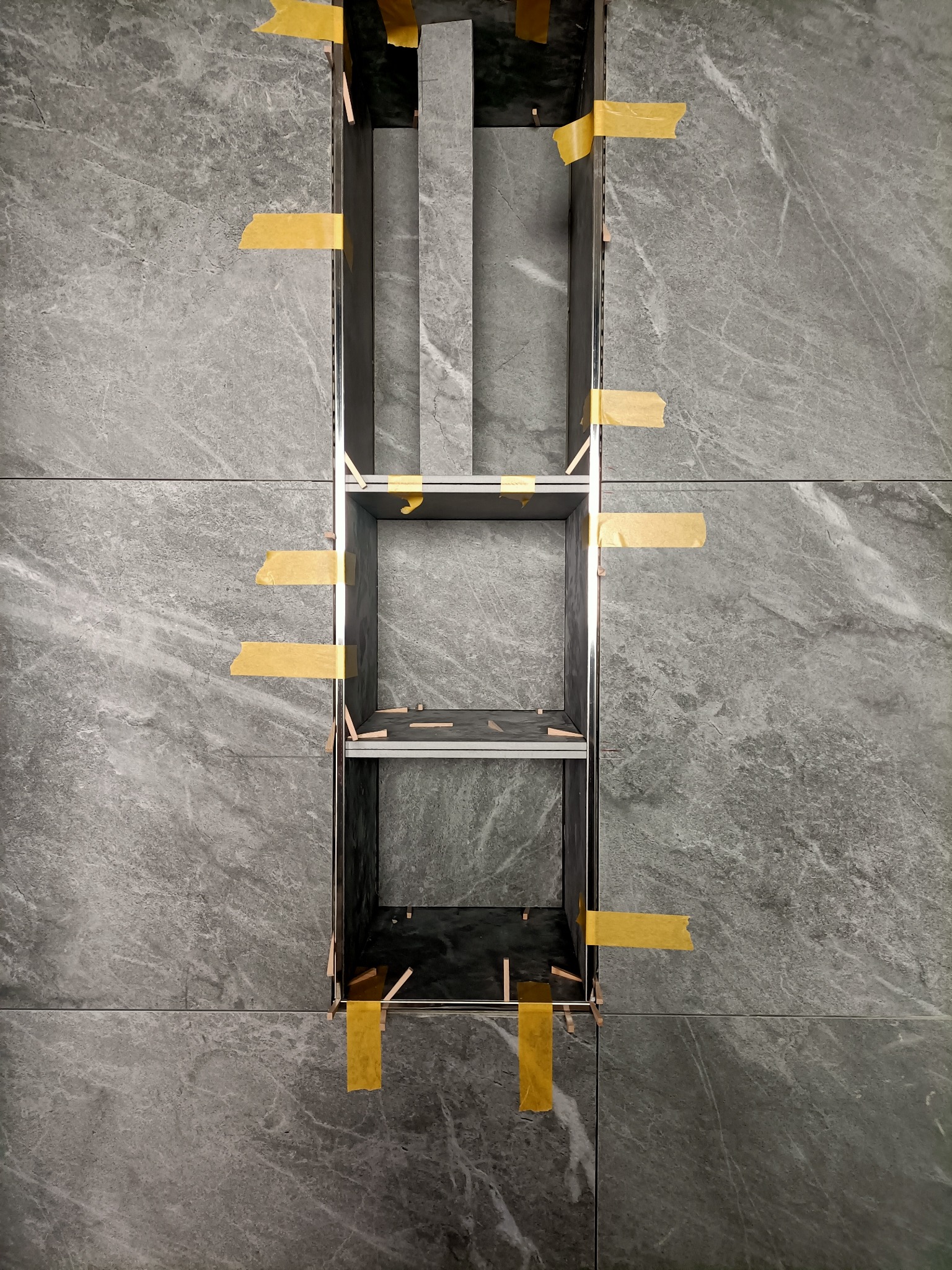 Wnęka w ścianie wyłożonej szarymi płytkami imitującymi marmur, z widocznymi elementami konstrukcyjnymi mocowanymi taśmą malarską i klinami dystansowymi.