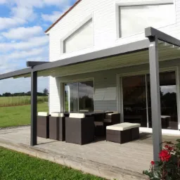 Nowoczesna pergola z regulowanym zadaszeniem, stojąca na drewnianym tarasie przy domu z dużymi przesuwnymi drzwiami, obok zestaw mebli ogrodowych z ciemnej plecionki.