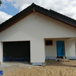 Jednopiętrowy dom z garażem w trakcie prac elewacyjnych, z widocznym ociepleniem fundamentów, zabezpieczone drzwi folią ochronną, piasek wokół budynku.