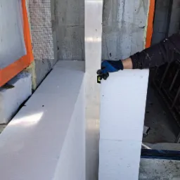 Ocieplanie elewacji styropianem: pracownik w rękawicach ustawia poziom pionowy przy pomocy poziomicy na aluminiowej łacie, widoczna siatka zbrojeniowa i surowy beton.