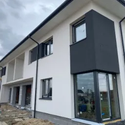 Nowoczesny dom dwukondygnacyjny z częściowo wykończoną elewacją: biały tynk kontrastuje z ciemnoszarym fragmentem przy oknie, widoczne ślady prac budowlanych i narzędzia na pierwszym planie.