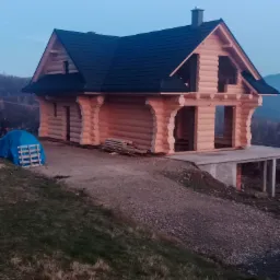 Surowy drewniany dom w trakcie budowy z czarnym dachem, widoczna więźba dachowa i podłoże z betonu, w tle zalesione wzgórza w pochmurny dzień.